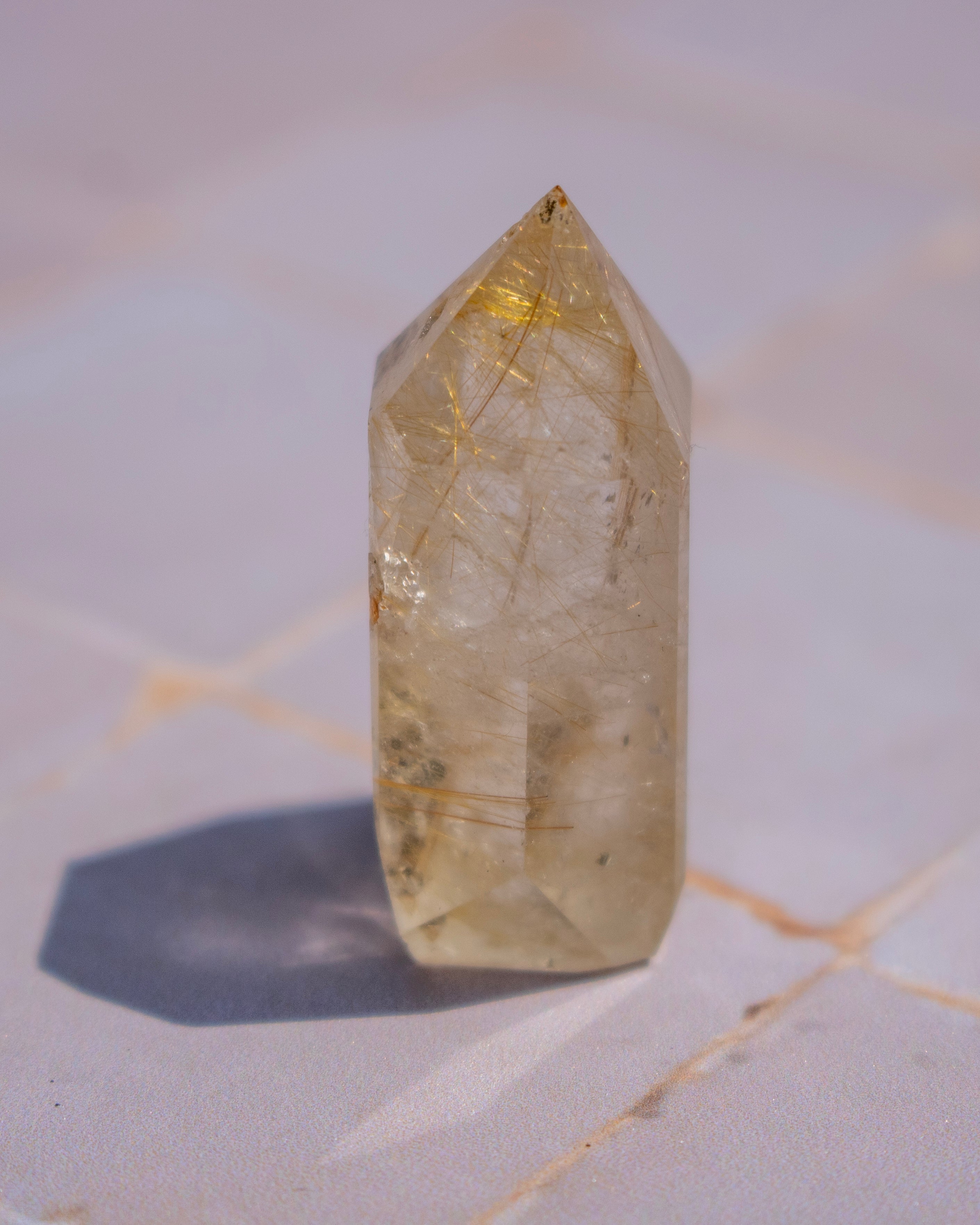 A natural clear quartz point crystal with prominent golden needle inclusions, standing upright on a light marble surface.