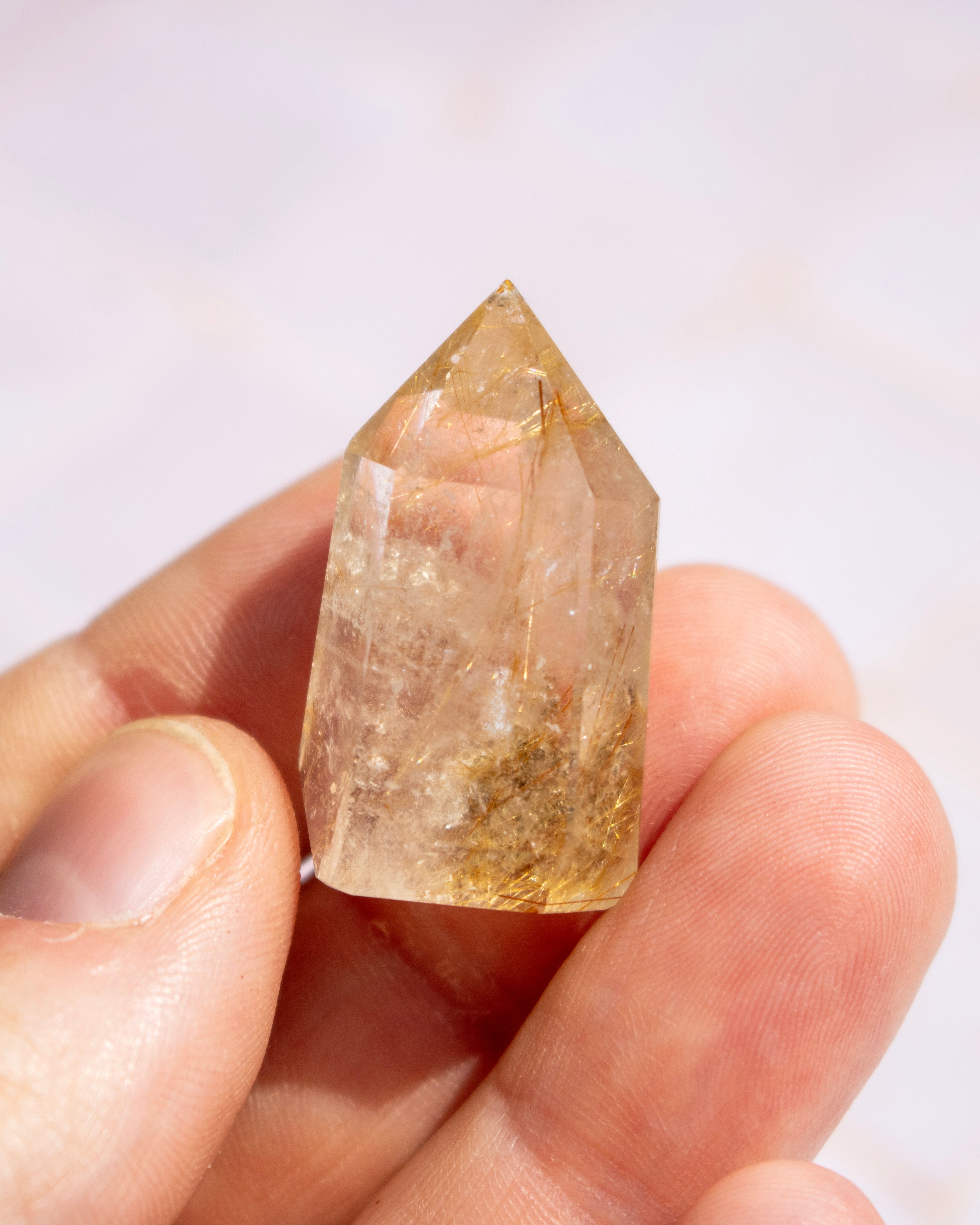 A small pointed clear quartz crystal with golden-brown inclusions, held between fingers against a soft light background.