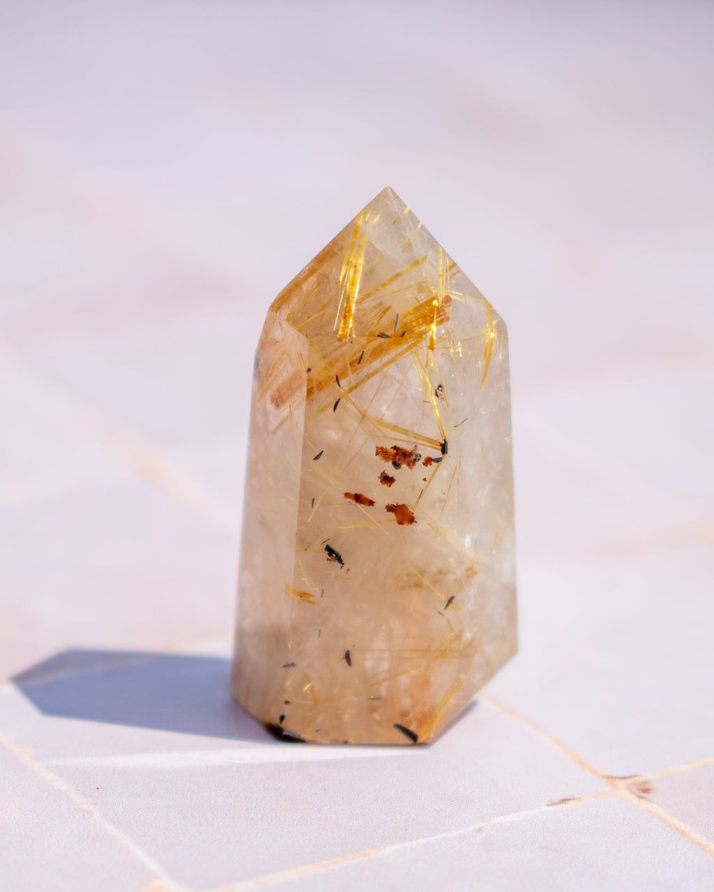 A natural quartz crystal point featuring prominent golden needle inclusions and small brownish inclusions, standing upright on a light tiled surface.
