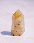 A natural quartz crystal point featuring prominent golden needle inclusions and small brownish inclusions, standing upright on a light tiled surface.