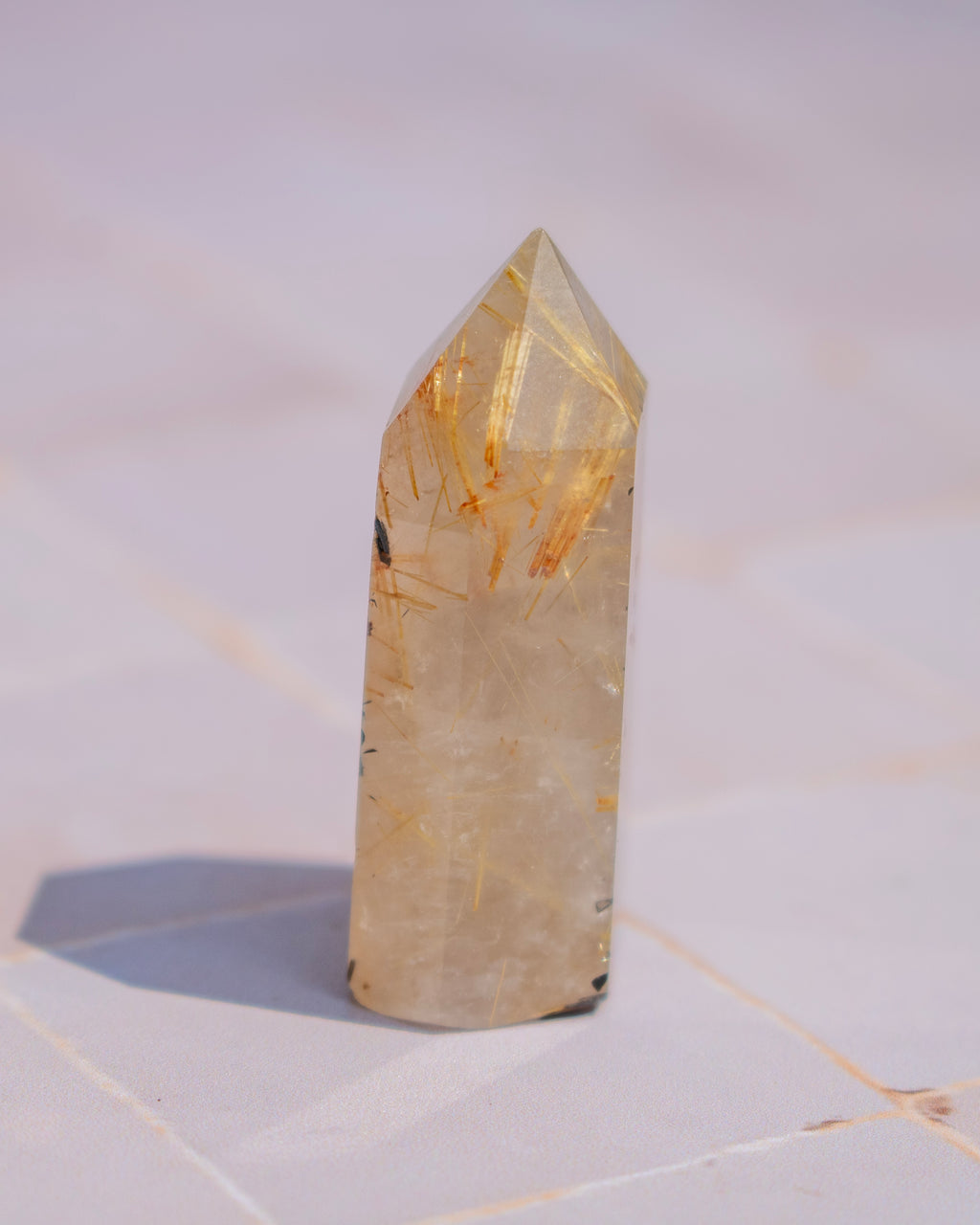 A pointed clear quartz crystal tower with prominent golden and orange metallic inclusions, standing on a light-colored tiled surface.