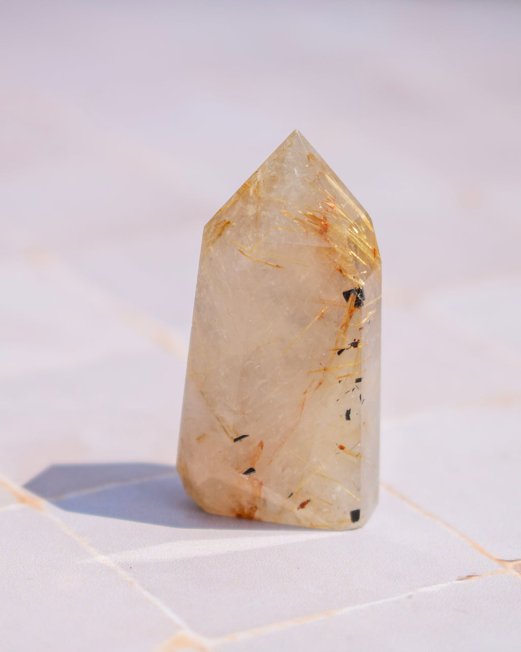 A pointed clear quartz crystal tower with golden and black inclusions, standing on a light tiled surface.