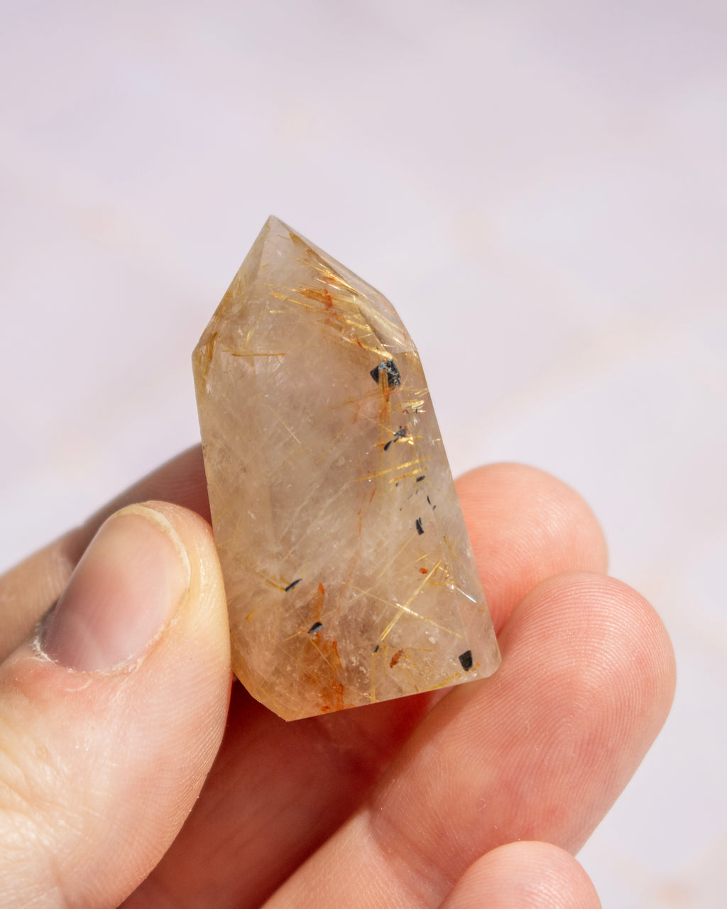 A hand holding a pointed crystal mineral specimen with a translucent pale beige body and visible golden and dark inclusions, against a soft light background.