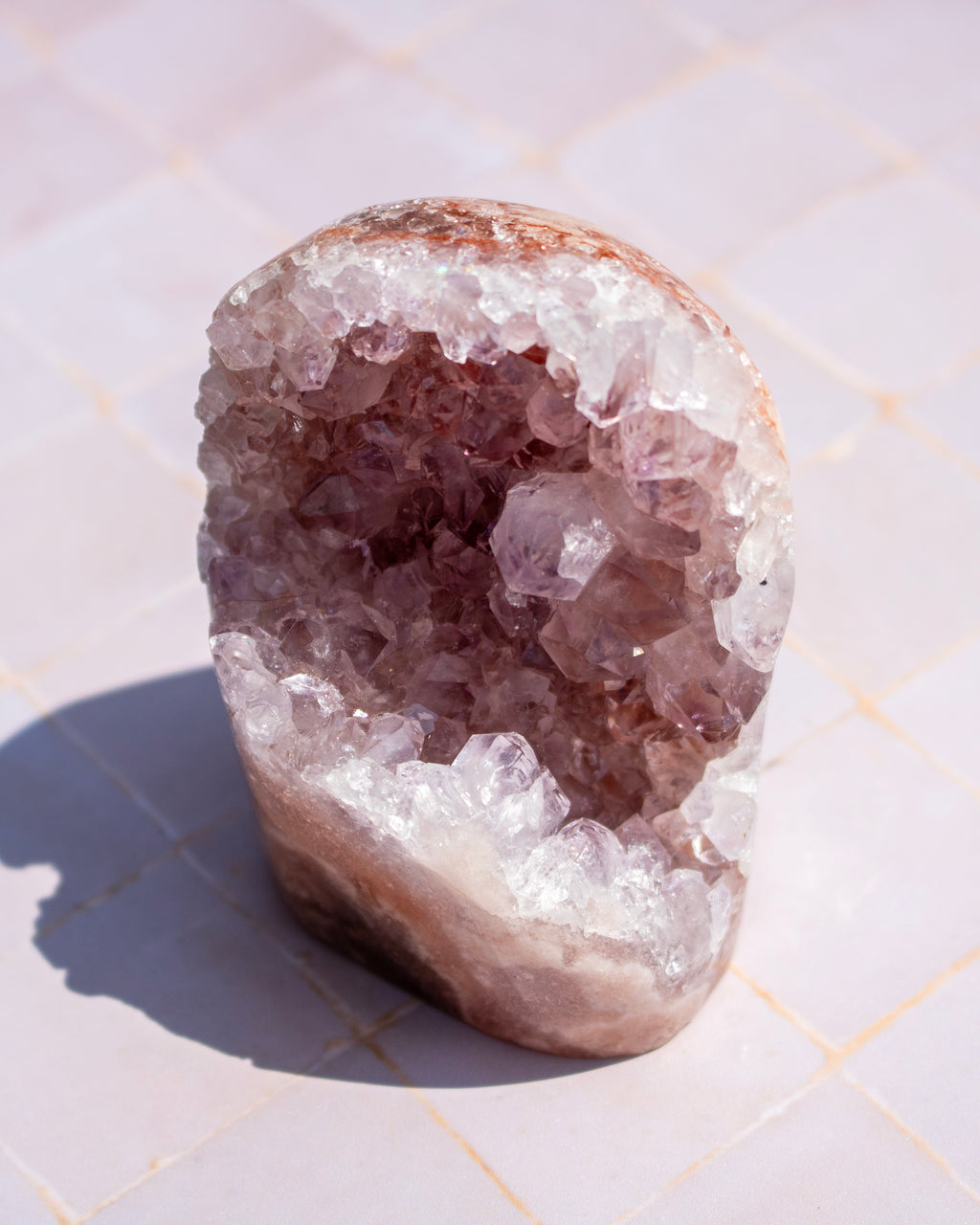 A raw amethyst crystal geode specimen with a hollow interior filled with purple crystals, photographed on a light tiled surface in direct sunlight.
