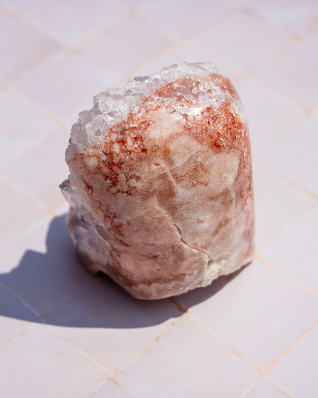 A rough, natural crystal or stone specimen with a mix of translucent white areas and warm peachy-brown mineral deposits, standing on a light tiled surface.