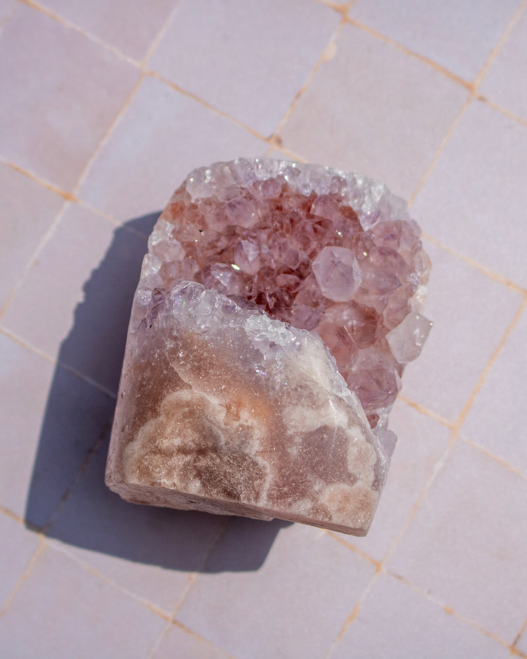 A raw amethyst crystal cluster with pinkish-purple quartz points and a rough, textured beige matrix base, photographed on a light tiled surface.
