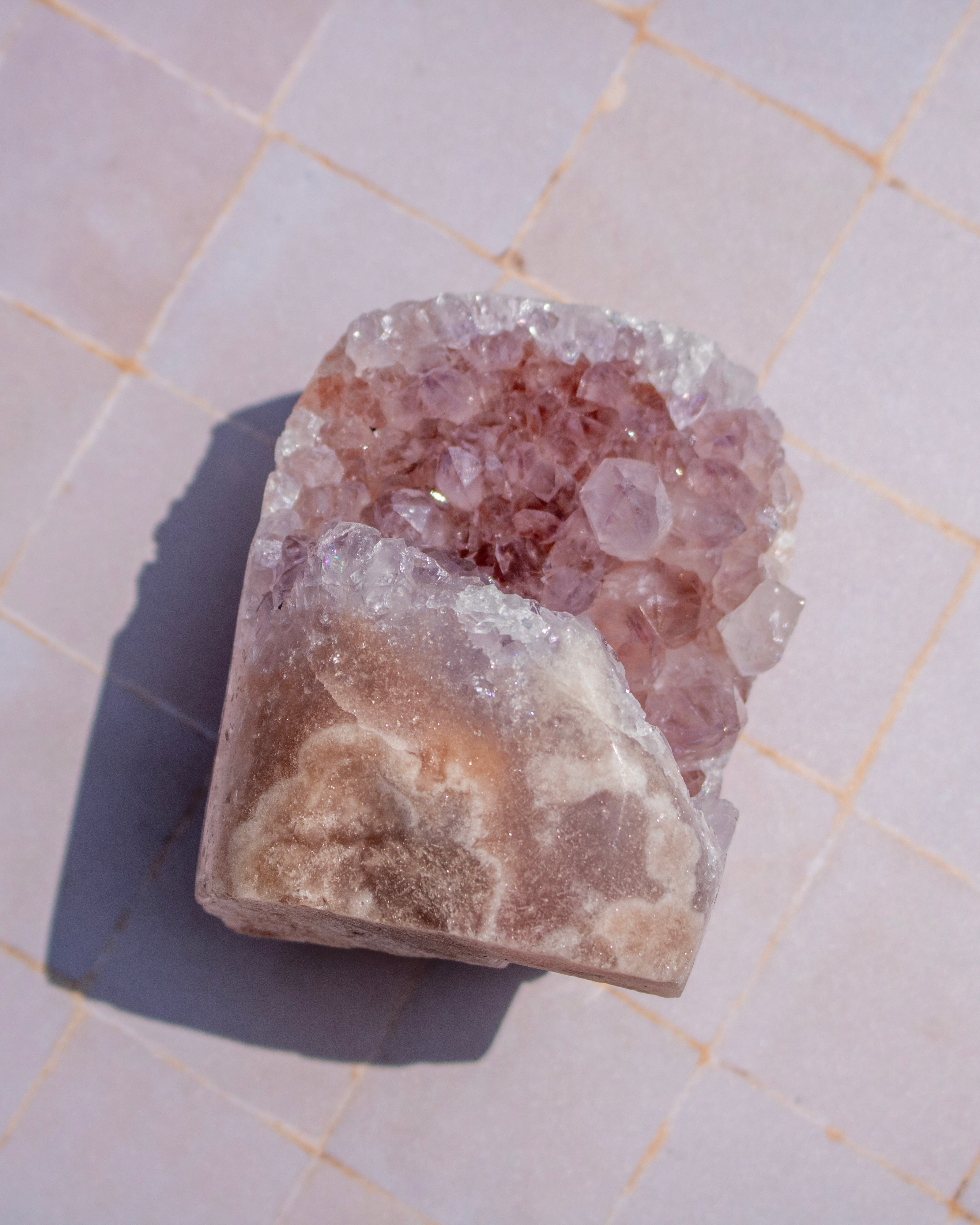 A raw amethyst crystal cluster with pinkish-purple quartz points and a rough, textured beige matrix base, photographed on a light tiled surface.