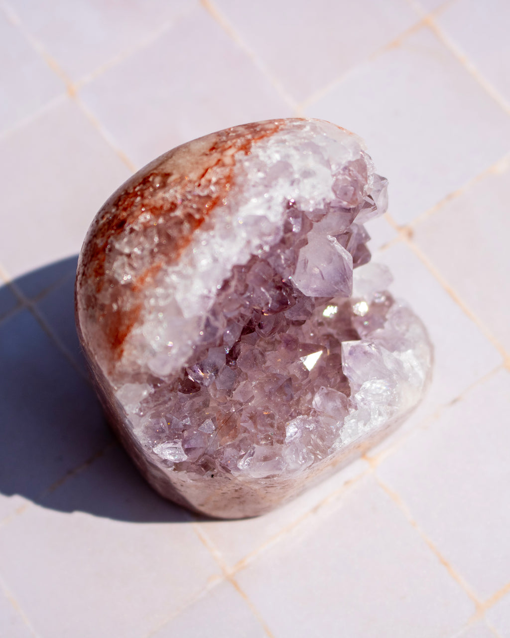 A polished natural crystal geode specimen with a rough brown outer surface and a clear lavender-purple crystal interior, photographed on a light tiled surface.