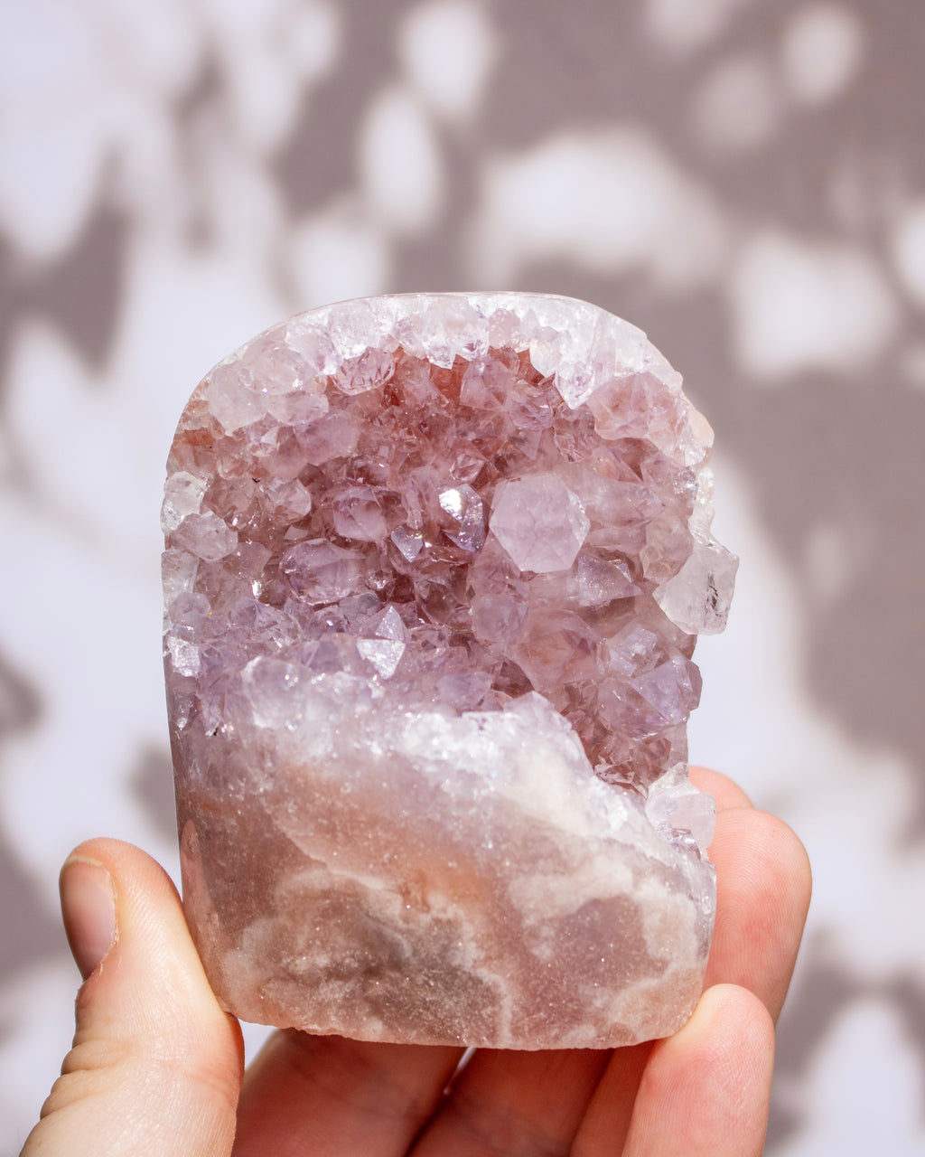 A hand holding a raw amethyst crystal cluster geode with light purple and pinkish-lavender crystalline formations against a soft-focus background with leaf shadows.