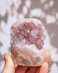 A hand holding a raw amethyst crystal cluster geode with light purple and pinkish-lavender crystalline formations against a soft-focus background with leaf shadows.