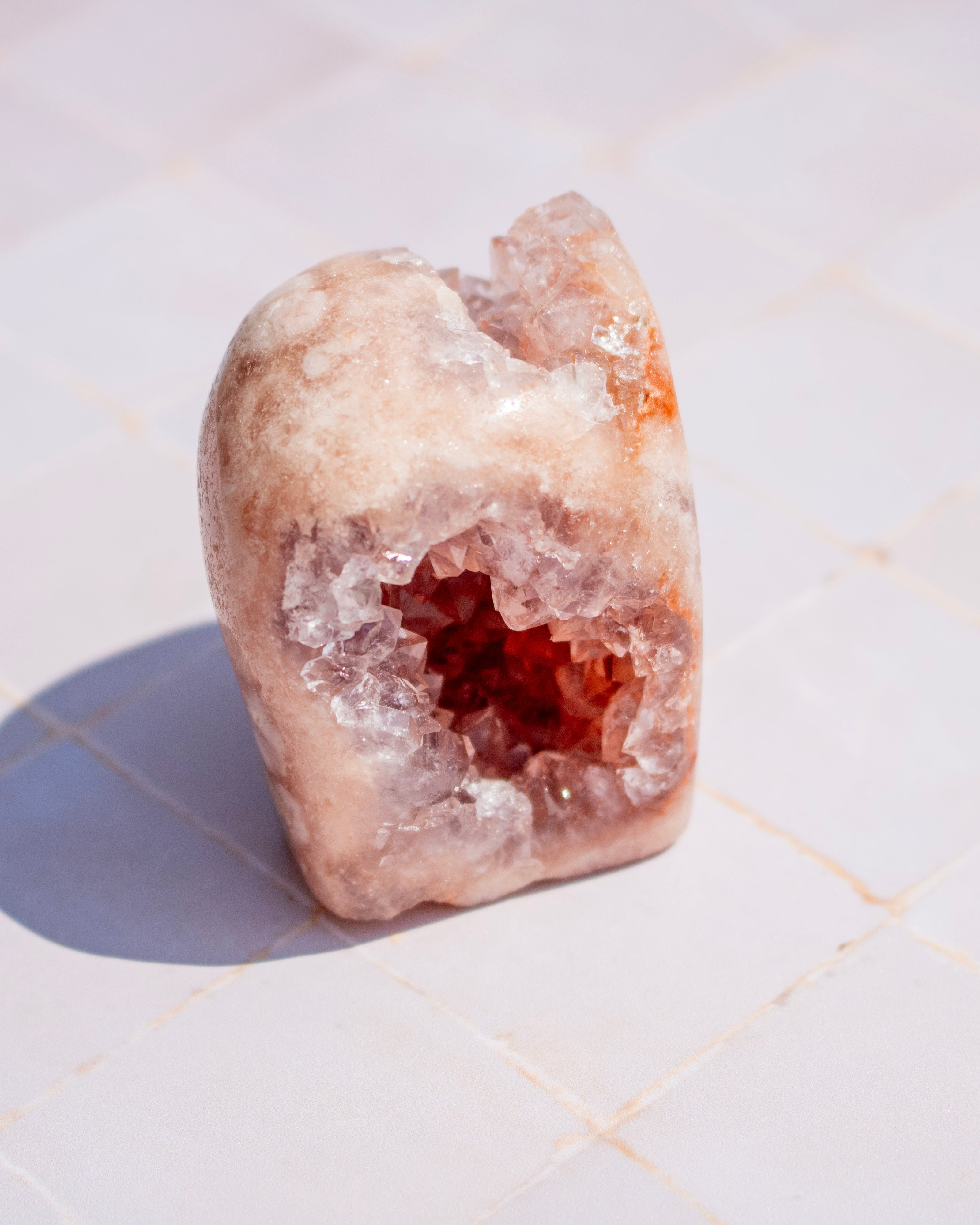 A raw crystal geode specimen with a rough, polished outer surface in pale peach and cream tones, featuring a hollow interior filled with clear quartz crystal clusters and a warm reddish-brown center, photographed on a light tiled surface.