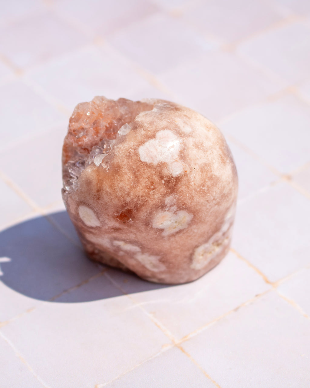 A rough, rounded peach-colored crystal cluster or stone with white crystalline inclusions, resting on a light tiled surface under bright sunlight.