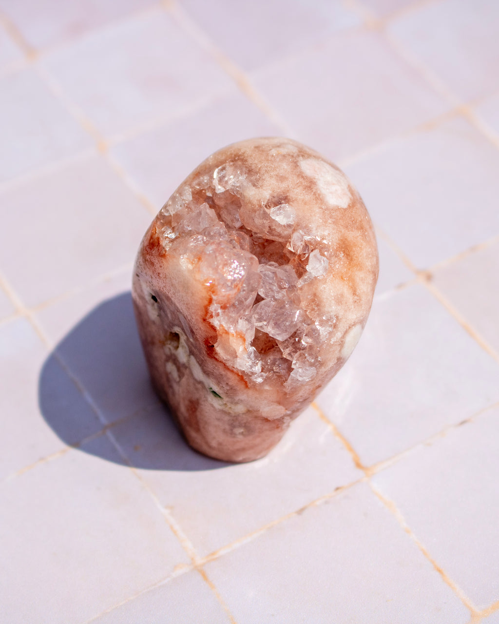 A polished peach-colored crystal stone with natural rough crystalline textures, standing upright on a light beige tiled surface under bright sunlight.