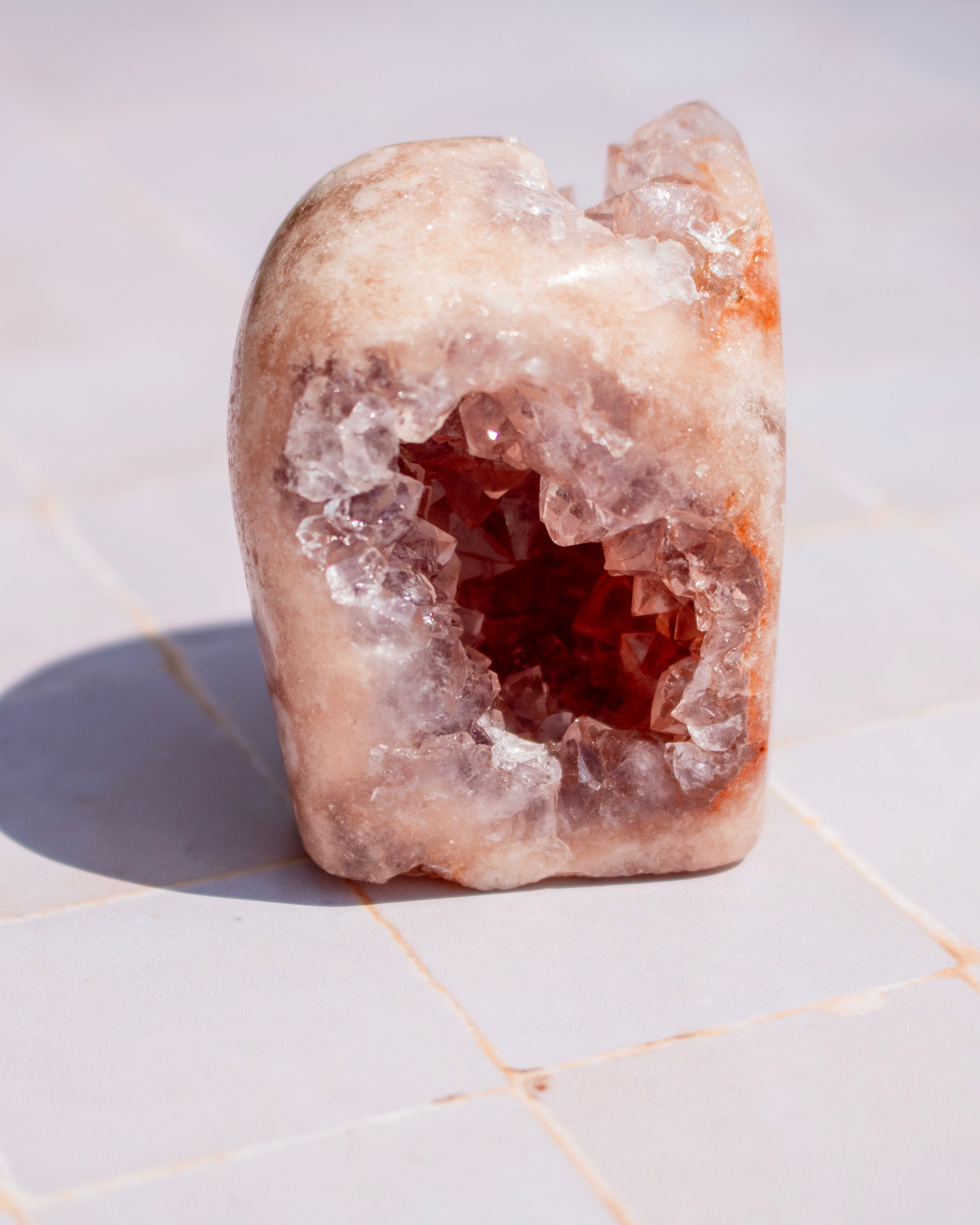 A raw crystal geode specimen with a pale peach and pink outer matrix and a deep reddish-brown cavity, standing upright on a light tiled surface.
