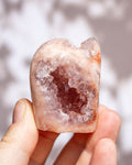 A hand holding a small raw crystal geode specimen with a pale pink to peach outer layer and a darker pink crystal cavity inside, against a softly blurred light gray background.