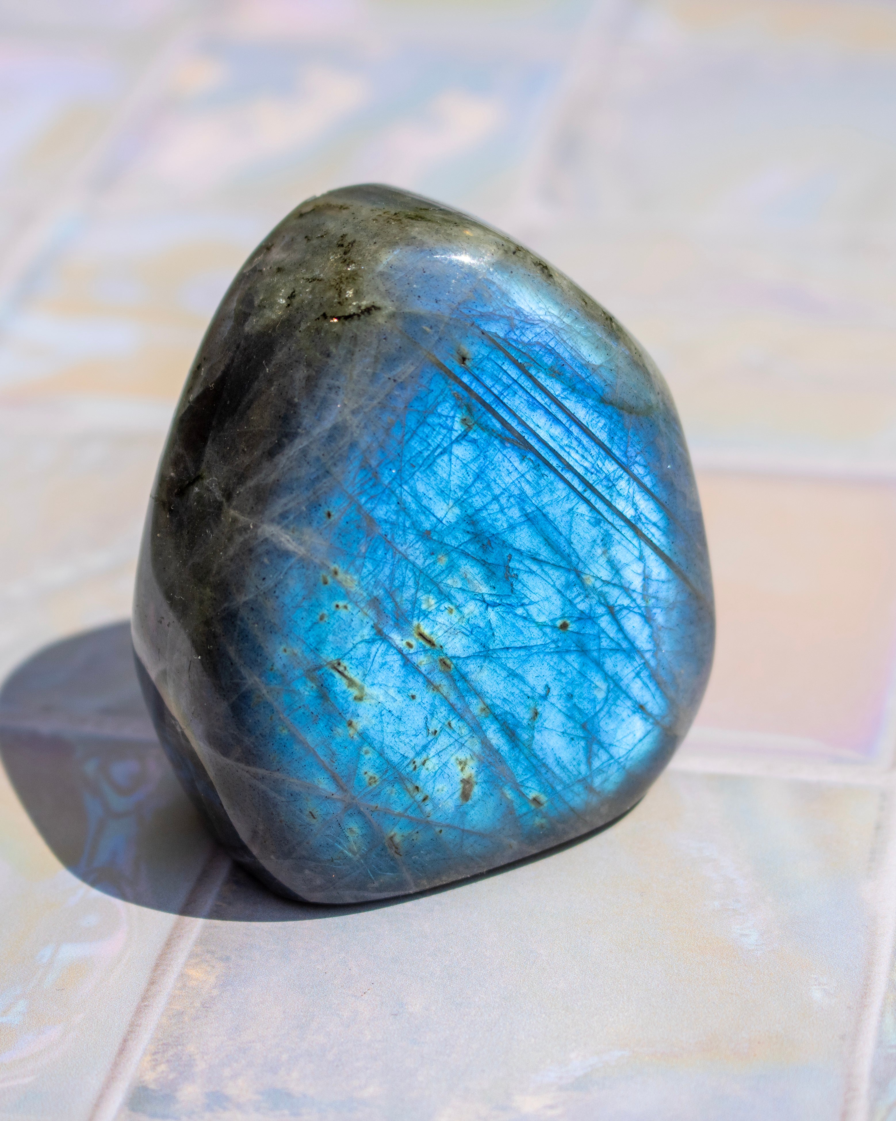 A polished tumbled labradorite stone with a shimmering blue flash and natural inclusions, resting on a light-colored tiled surface.