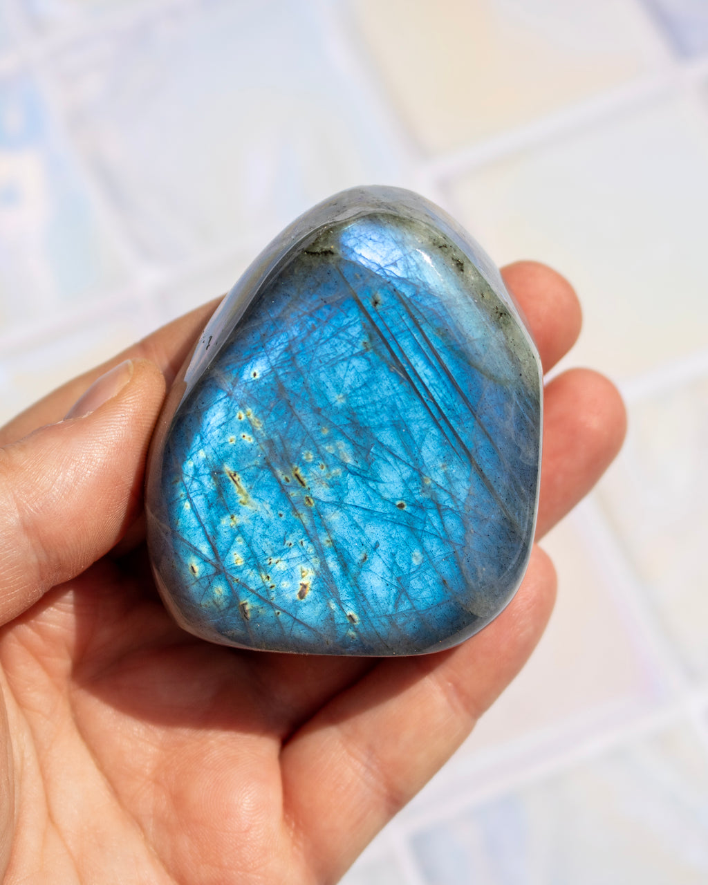 A polished blue labradorite stone held in a person's hand, showing a shimmering blue iridescent surface with fine crystalline lines and small gold flecks, against a softly blurred light background.