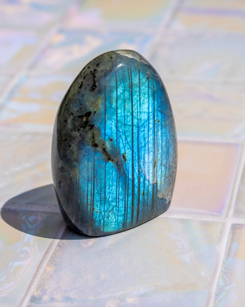 A polished labradorite crystal stone with a vibrant blue flash and dark gray base, standing upright on a light-colored tiled surface.