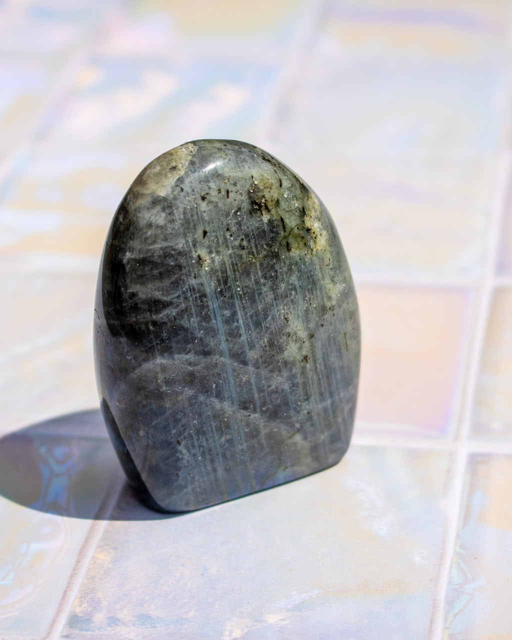 A polished labradorite crystal stone with a smooth, rounded teardrop shape, showcasing a dark gray body with natural inclusions and a blue-gray flash, standing on a light-colored tiled surface.