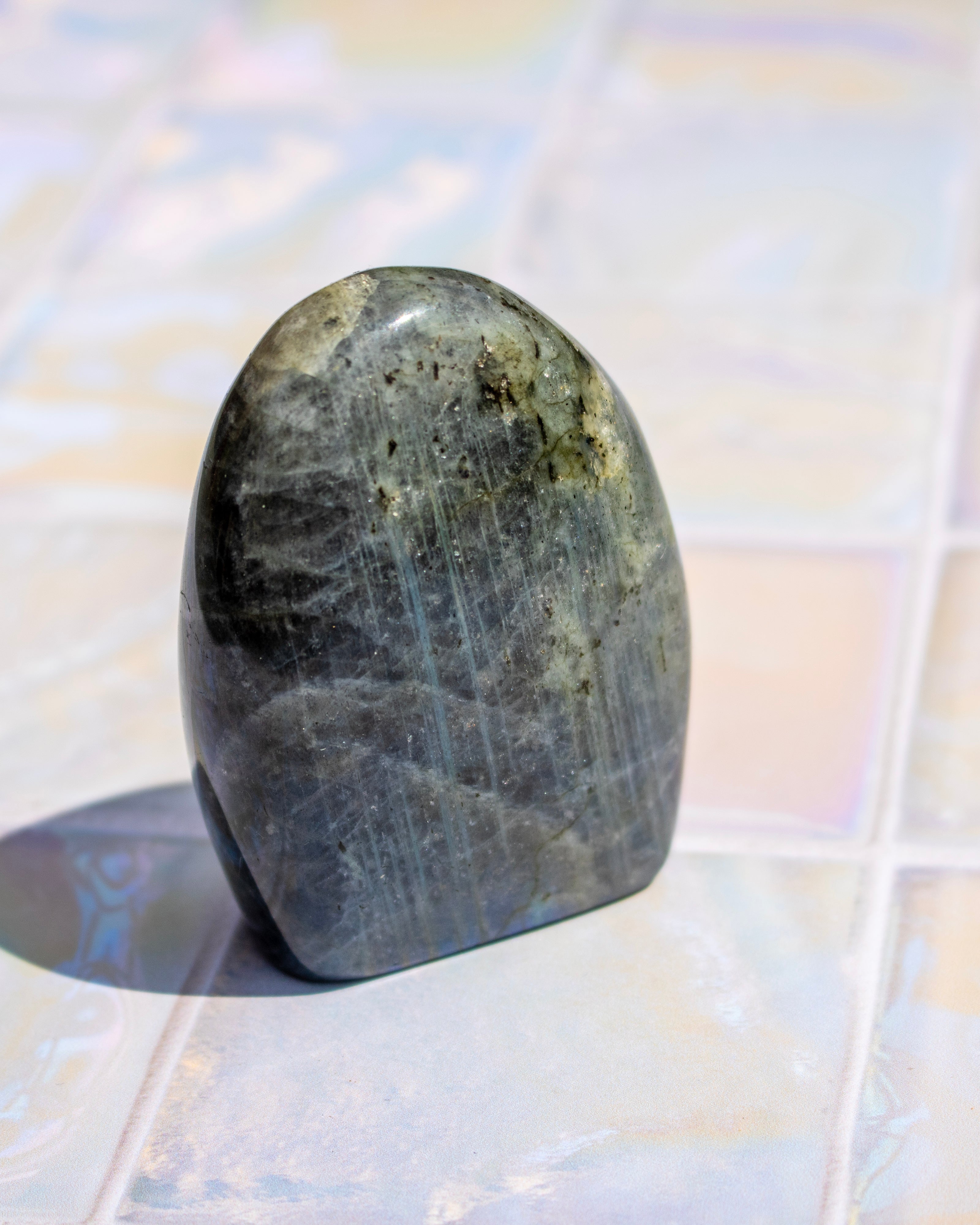 A polished labradorite crystal stone with a smooth, rounded teardrop shape, showcasing a dark gray body with natural inclusions and a blue-gray flash, standing on a light-colored tiled surface.