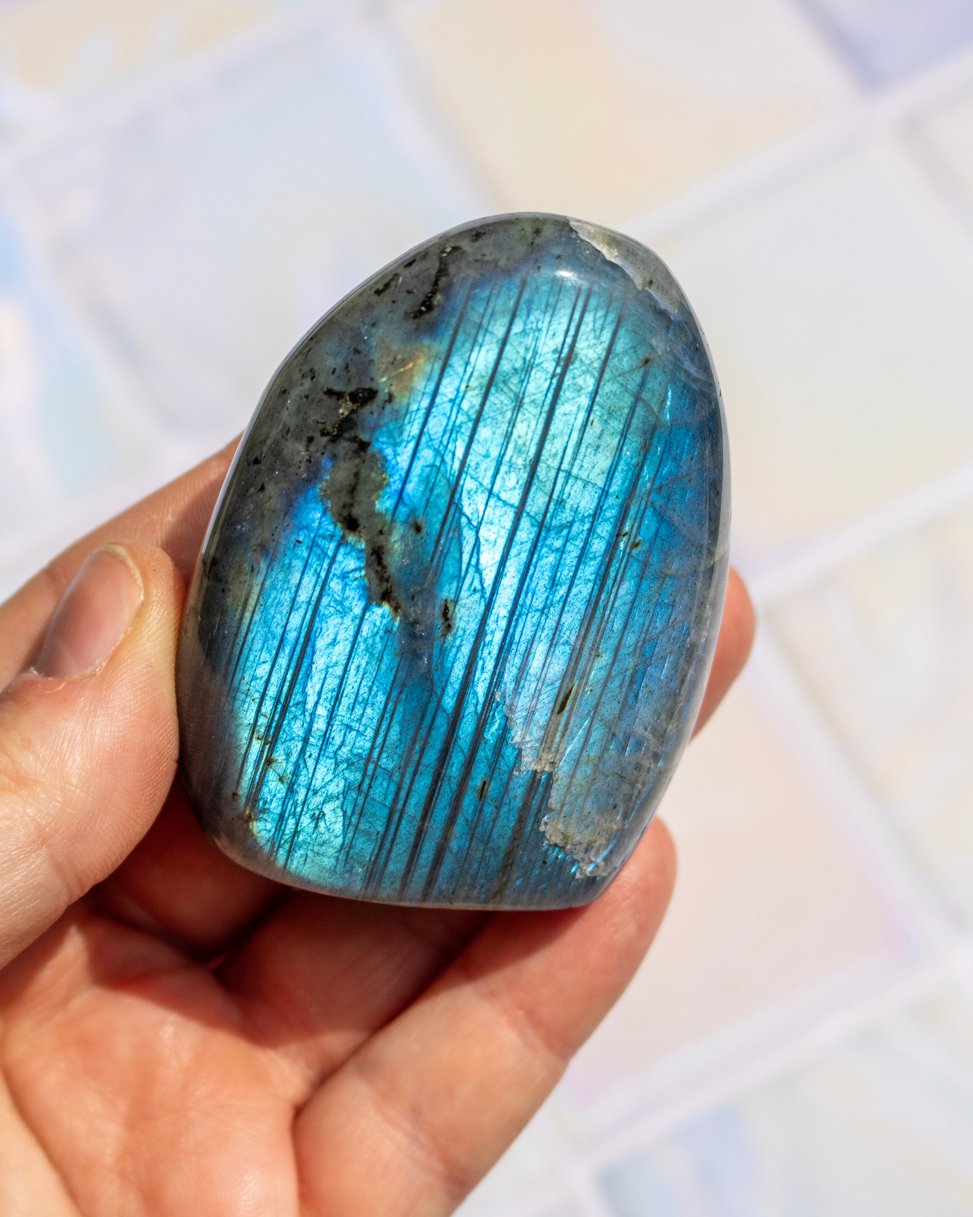 A polished labradorite crystal held in a person's hand, showing a shimmering blue and green iridescent surface with dark striations, against a softly blurred light background.
