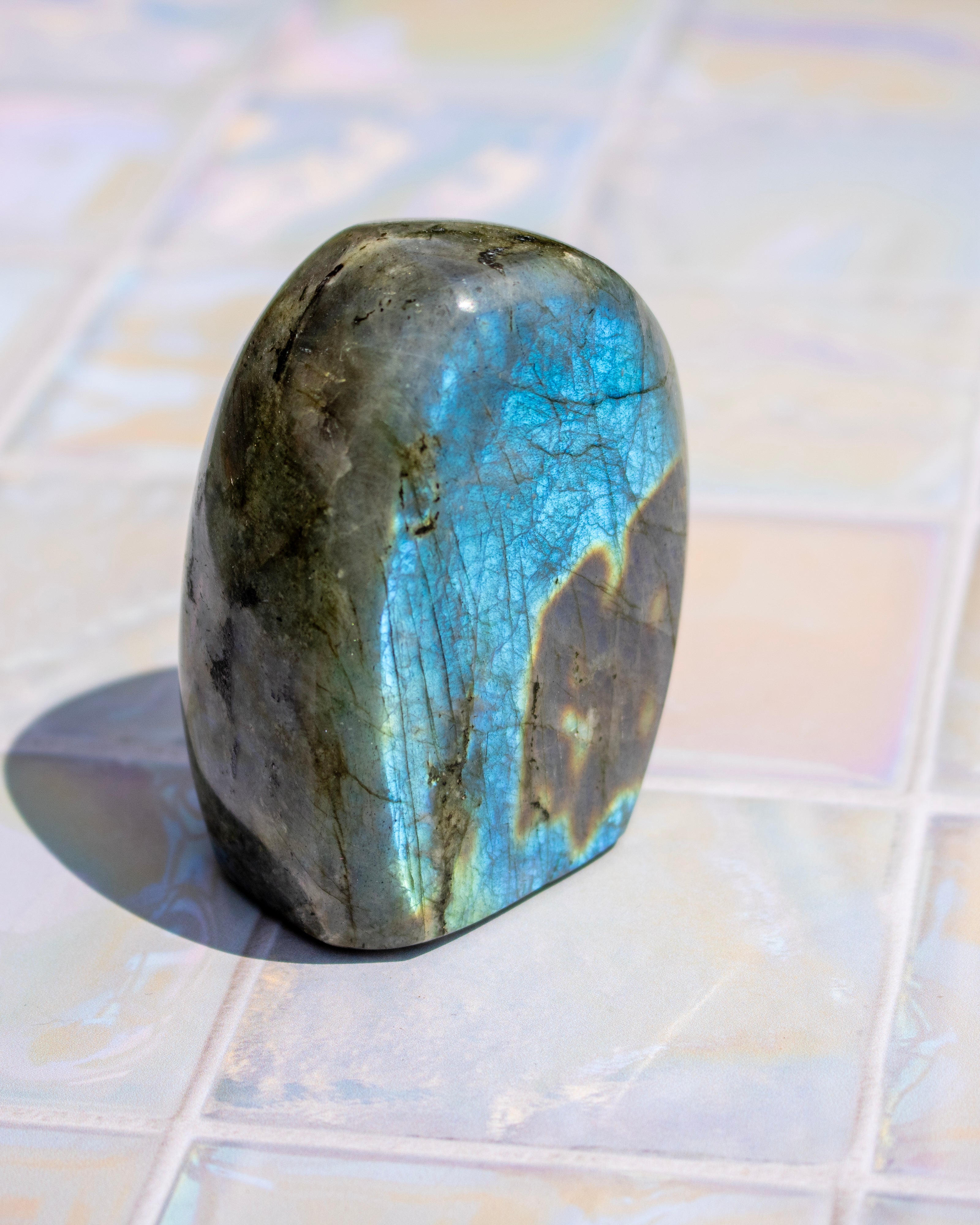 A polished labradorite crystal stone with a blue iridescent sheen and dark gray base, standing upright on a light-colored tiled surface.