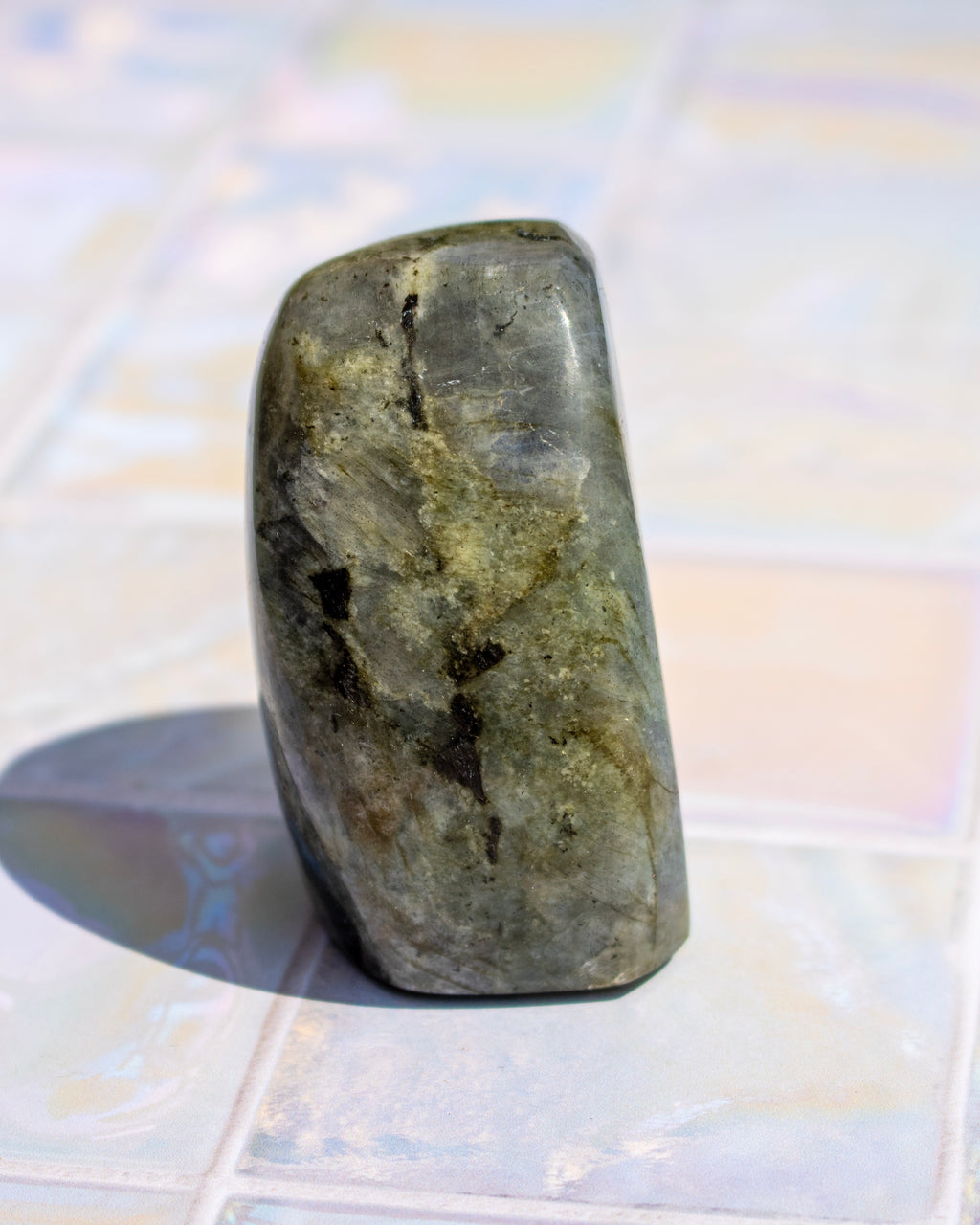 A polished natural stone or crystal with a rounded, tapered shape, standing upright on a light-colored tiled surface. The stone has a mottled texture of olive green, gray, and dark brown/black inclusions, casting a soft shadow to the left.