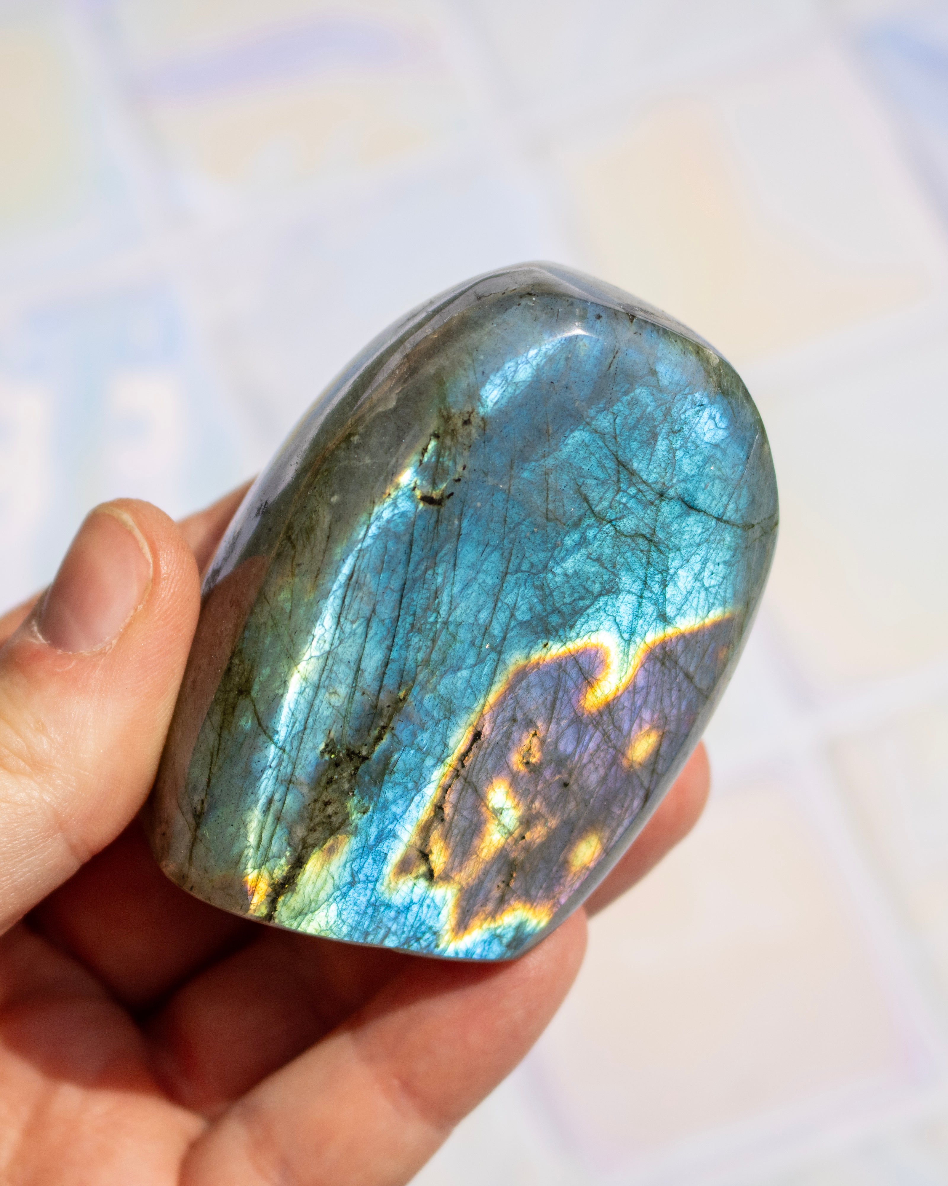 A hand holding a polished labradorite stone that displays a blue-green base color with a shimmering gold and purple flash.
