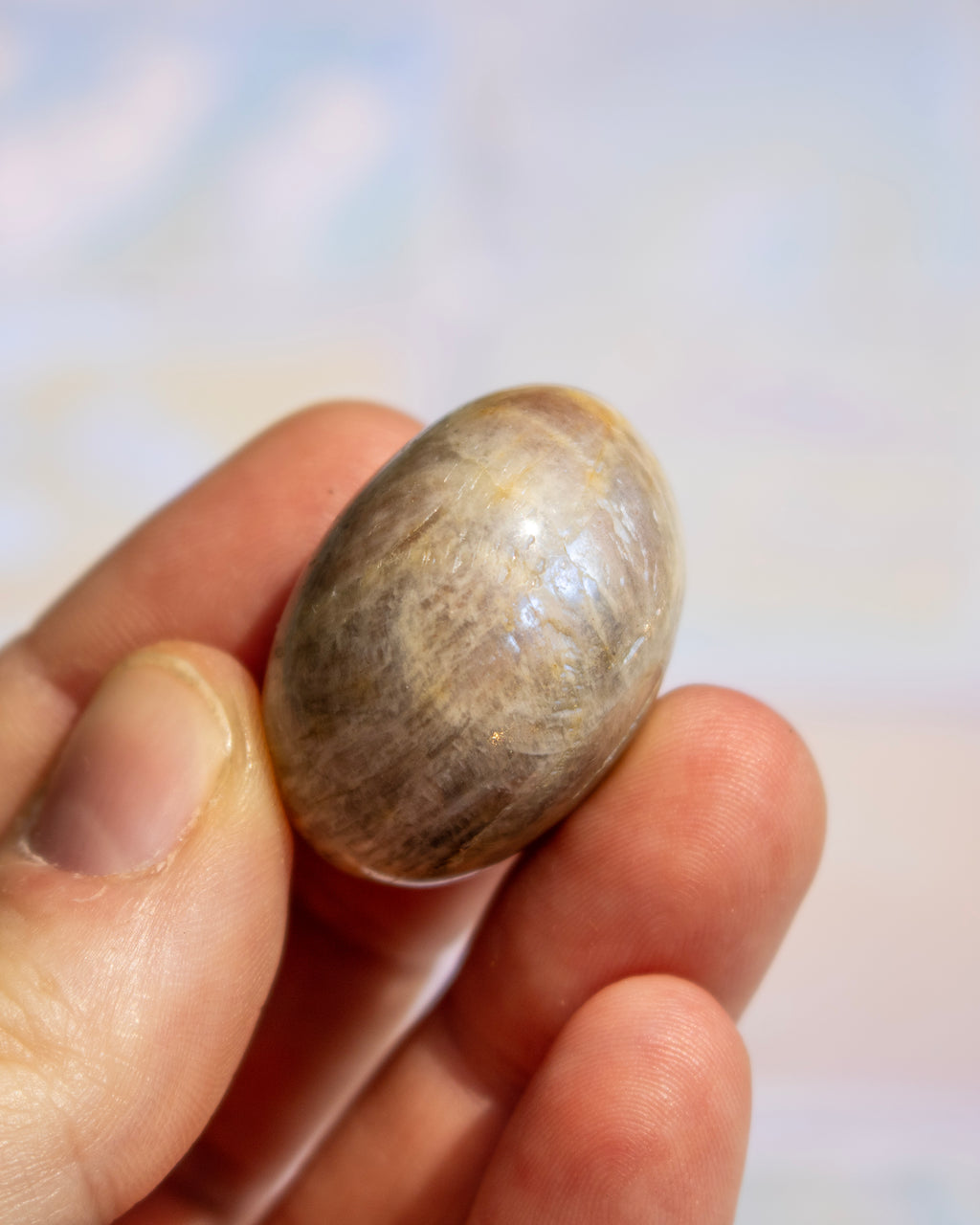 A small, smooth, oval-shaped stone or bead with natural brown and beige marbled patterns, held between fingers against a soft, out-of-focus light background.