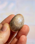 A small, smooth, oval-shaped stone or bead with natural brown and beige marbled patterns, held between fingers against a soft, out-of-focus light background.