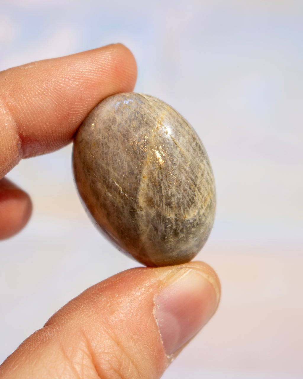 A close-up of a hand holding a smooth, polished oval stone with natural brown and beige marbling against a soft, out-of-focus light background.