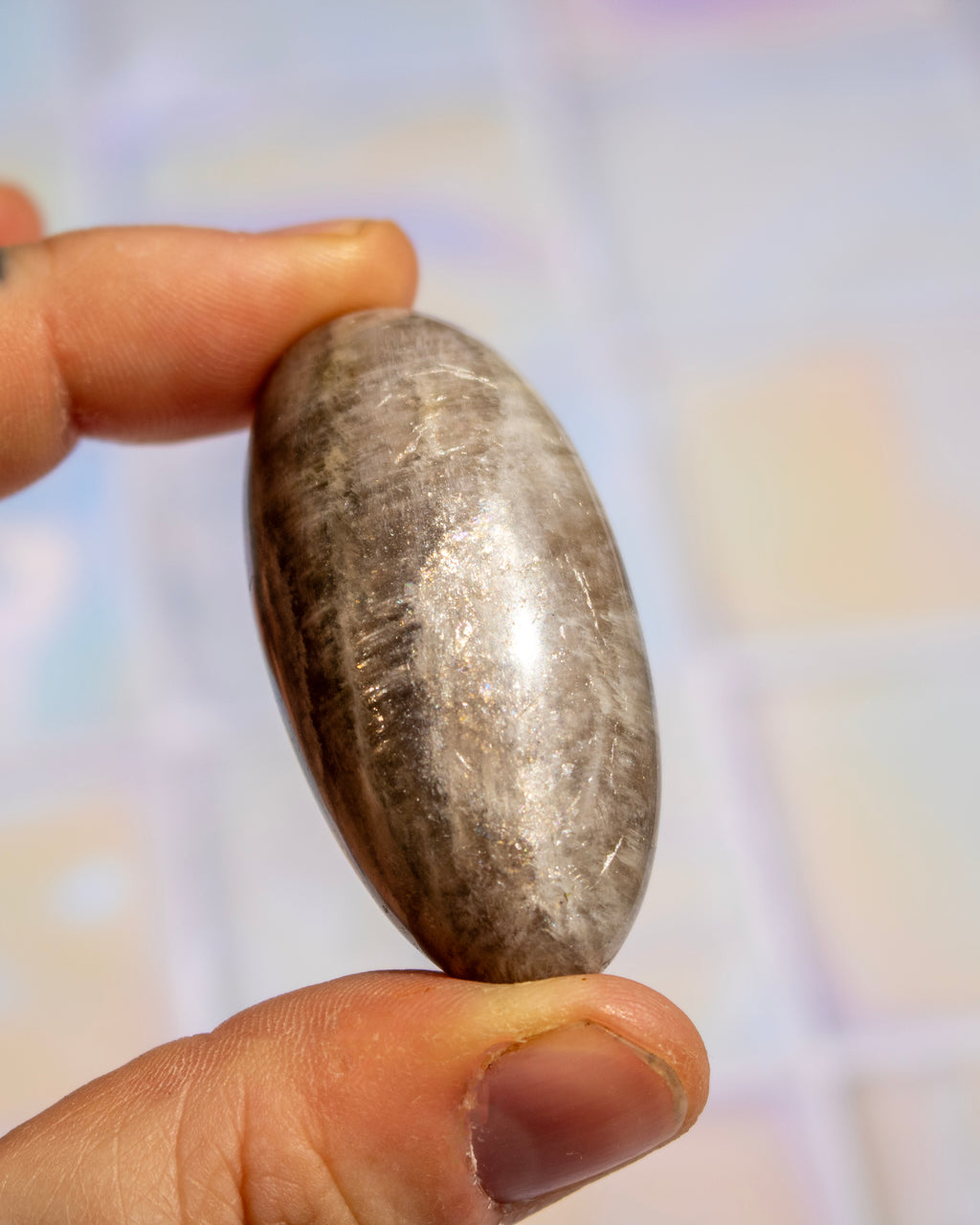 A hand holding a polished, smooth oval stone with a metallic silver-gray sheen and dark inclusions, set against a softly blurred pastel background.