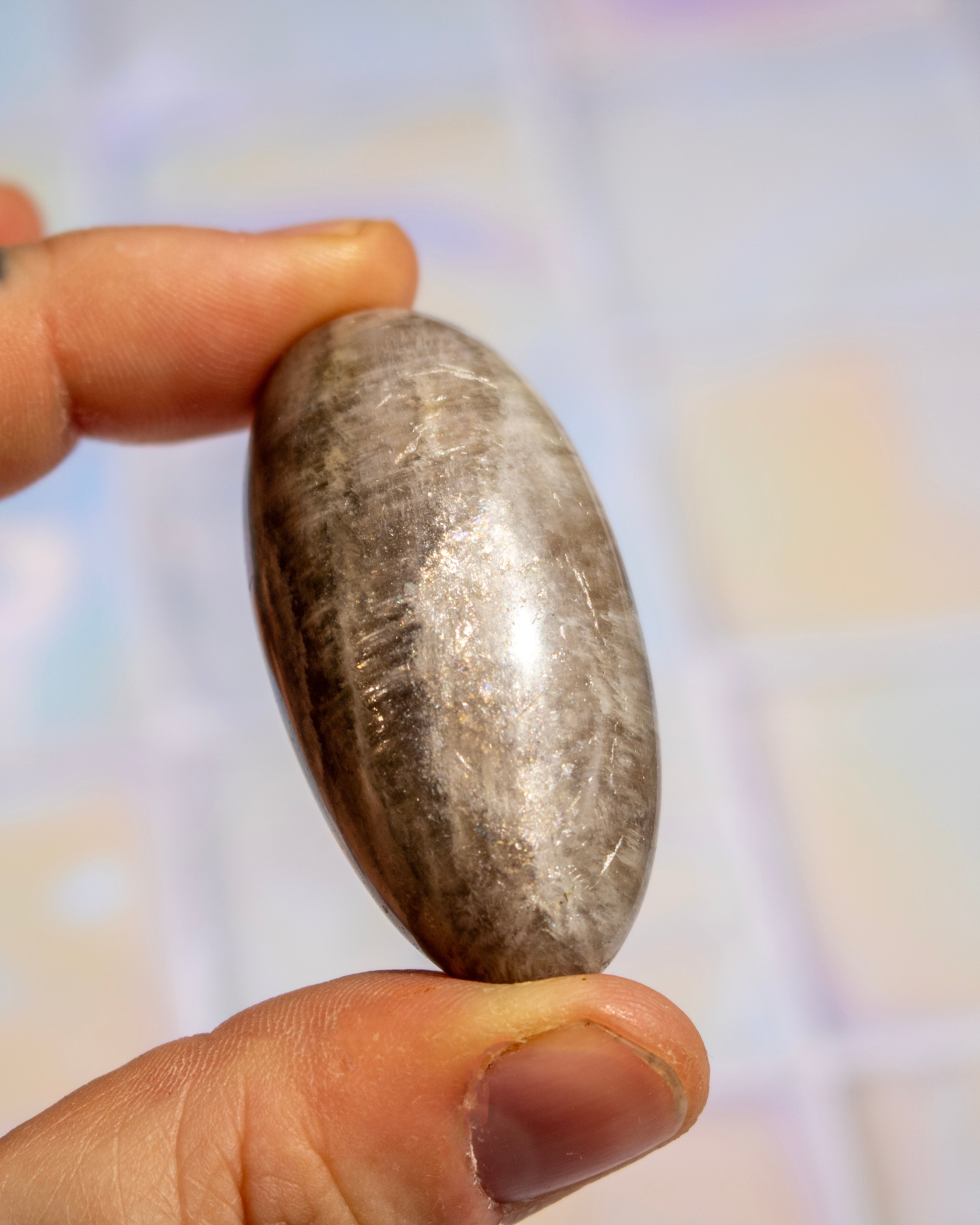 A hand holding a polished, smooth oval stone with a metallic silver-gray sheen and dark inclusions, set against a softly blurred pastel background.