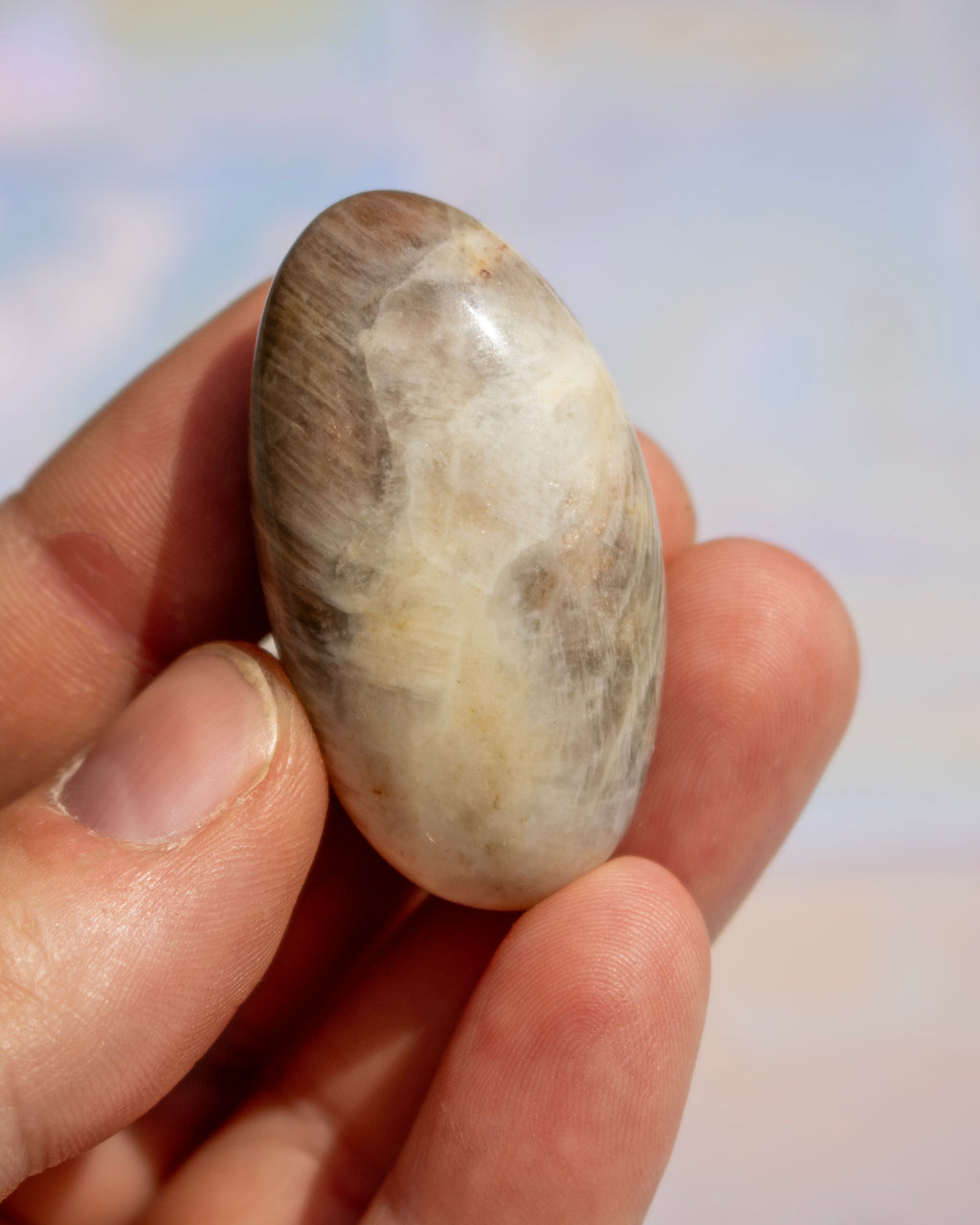 A hand holding a polished oval stone with natural beige, cream, and brown marbled patterns against a soft, out-of-focus light background.