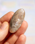 A polished oval stone with natural brown and beige marbling, held between a person's fingers against a soft, out-of-focus light background.