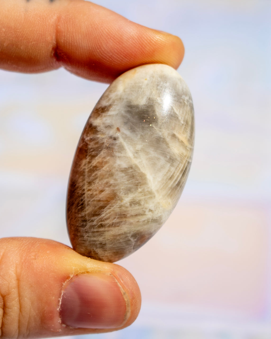 A polished oval stone with natural brown, beige, and gray marbling held between two fingers against a soft, out-of-focus pastel background.