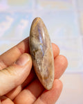A hand holding a polished oval stone with natural brown, beige, and gray marbling and subtle iridescent blue-purple sheen, against a softly blurred light background.