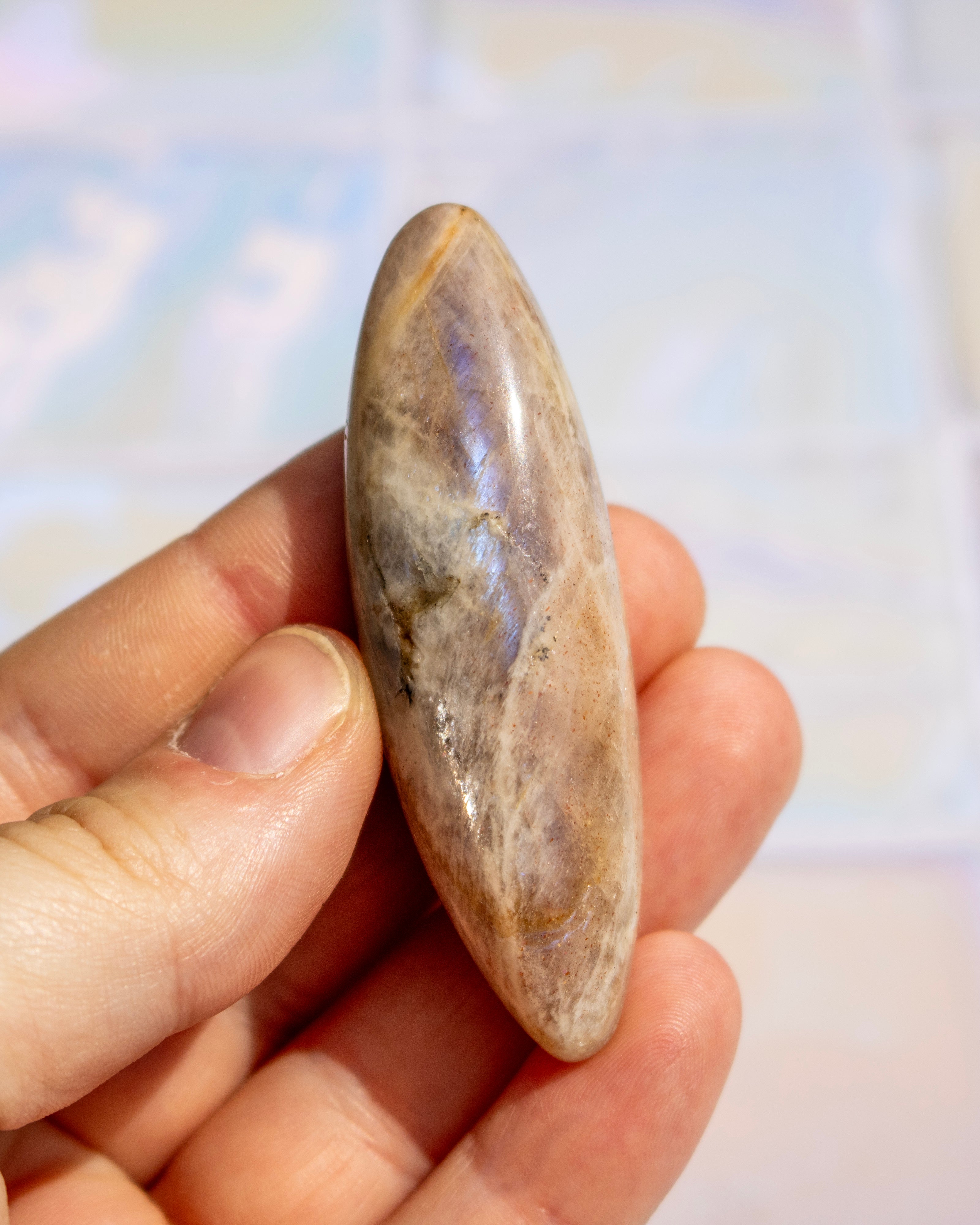 A hand holding a polished oval stone with natural brown, beige, and gray marbling and subtle iridescent blue-purple sheen, against a softly blurred light background.