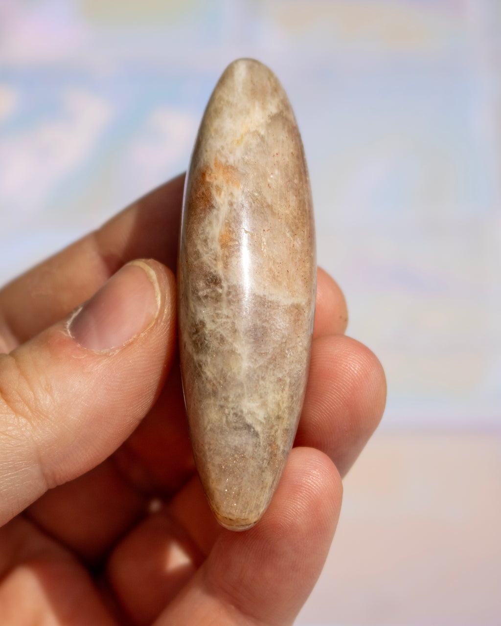 A hand holding a polished, teardrop-shaped stone with natural beige, brown, and cream marbling, set against a softly blurred pastel background.