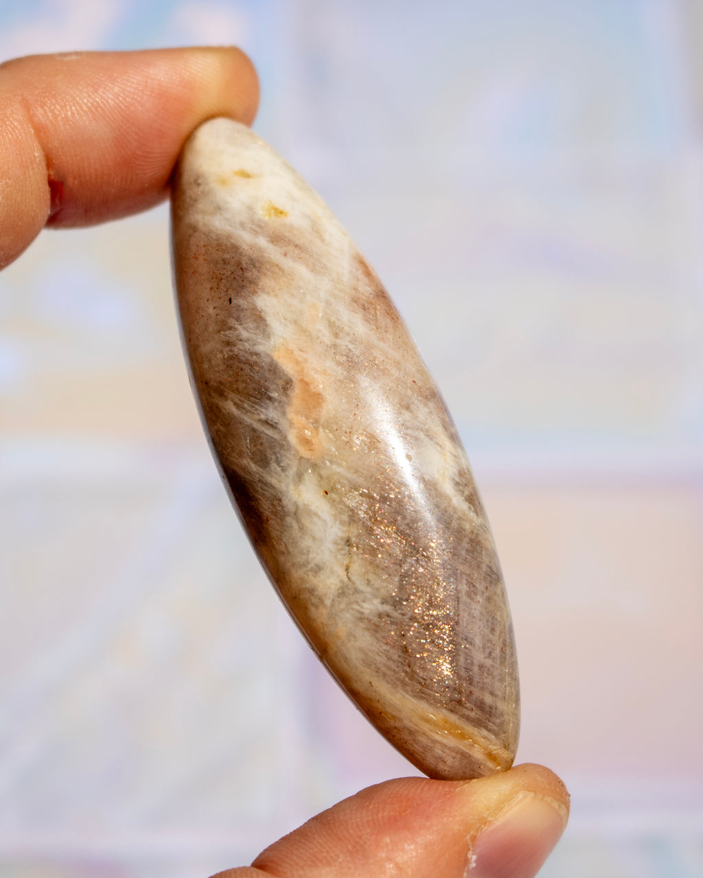 A hand holding a polished, teardrop-shaped stone cabochon with natural brown, beige, and white marbled patterns.