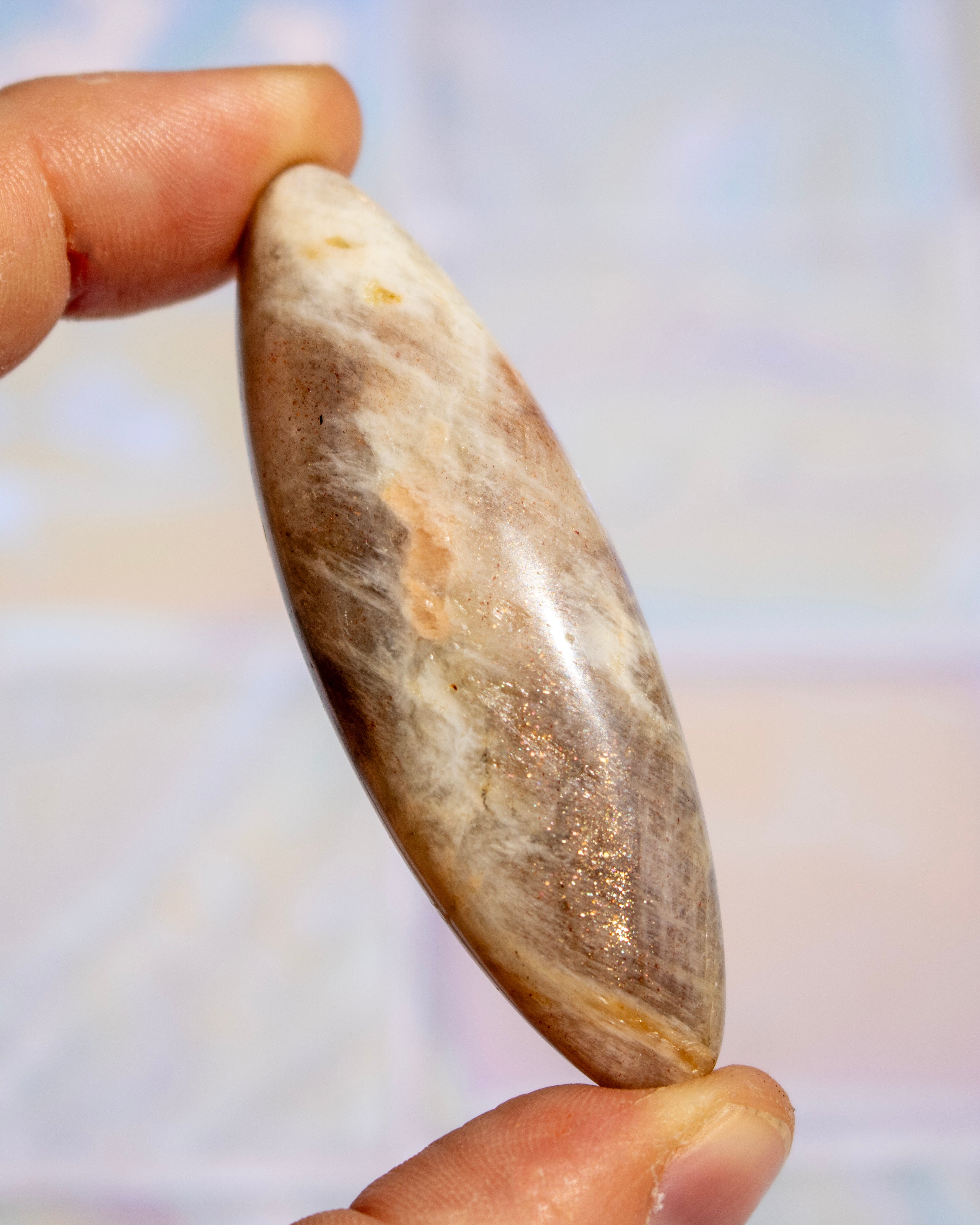 A hand holding a polished, teardrop-shaped stone cabochon with natural brown, beige, and white marbled patterns.
