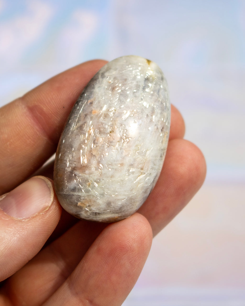 A hand holding a small, polished, oval-shaped clear crystal or glass bead with internal inclusions, set against a soft, out-of-focus light blue and white background.