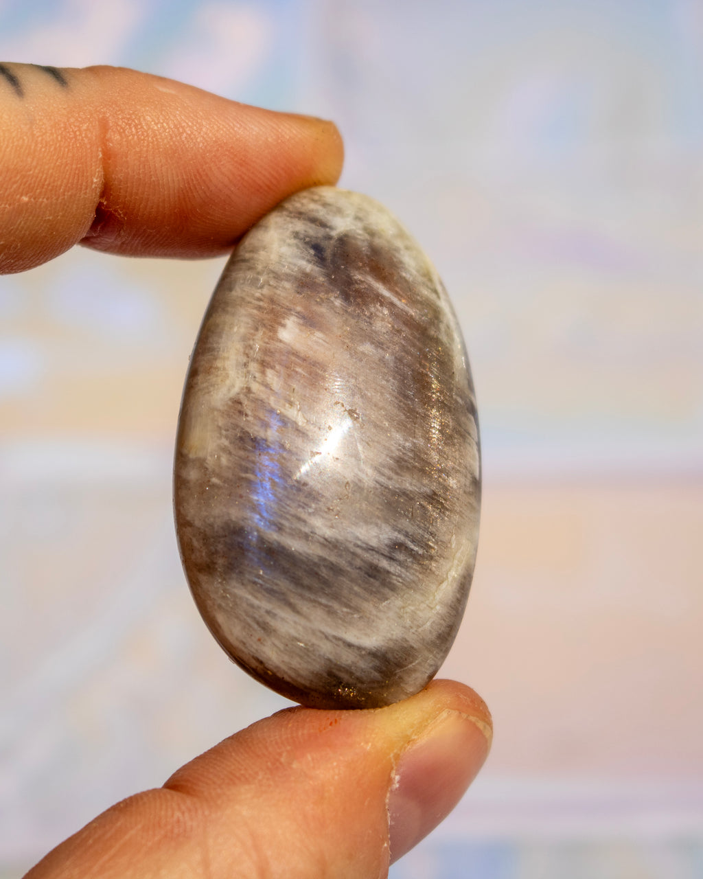 A polished, teardrop-shaped stone with natural gray, brown, and white marbled patterns, held between a person's fingers against a soft, out-of-focus background.