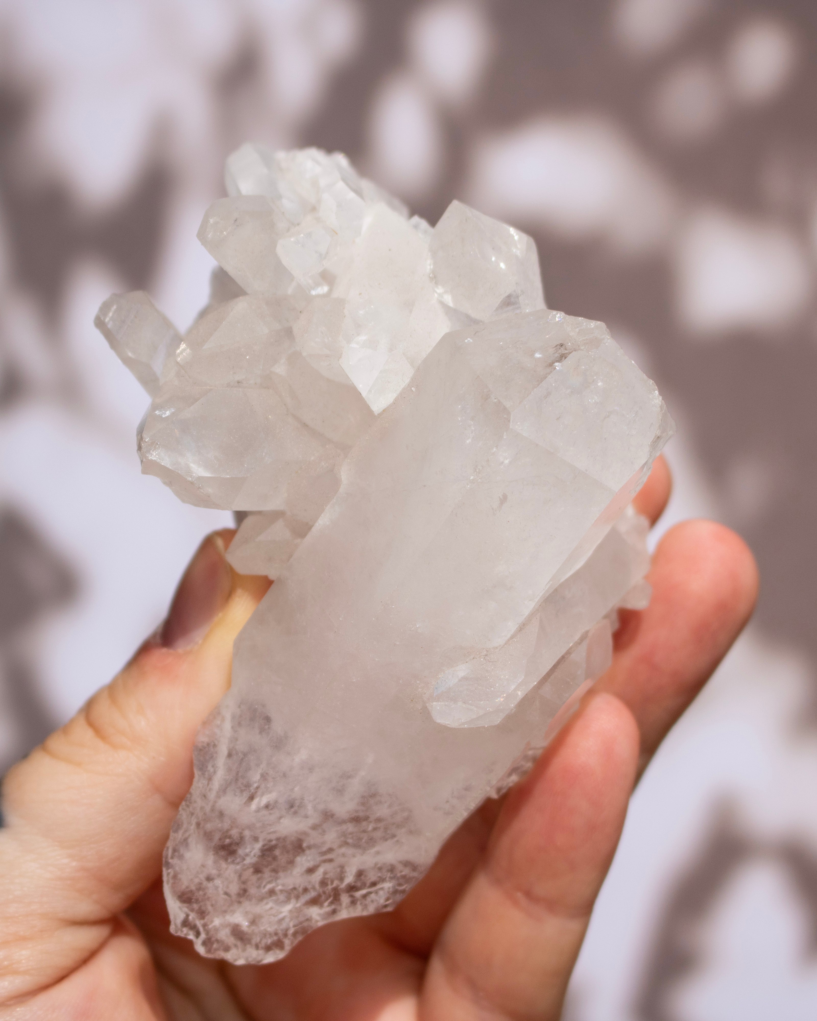 A hand holding a raw quartz crystal cluster with natural branching points and rough, unpolished edges, set against a soft-focus background with dappled light and shadow patterns.