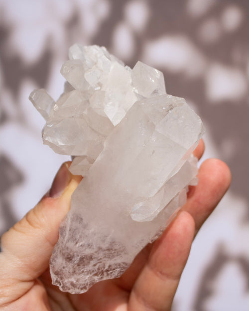 A hand holding a raw quartz crystal cluster with natural branching points and rough, unpolished edges, set against a soft-focus background with dappled light and shadow patterns.
