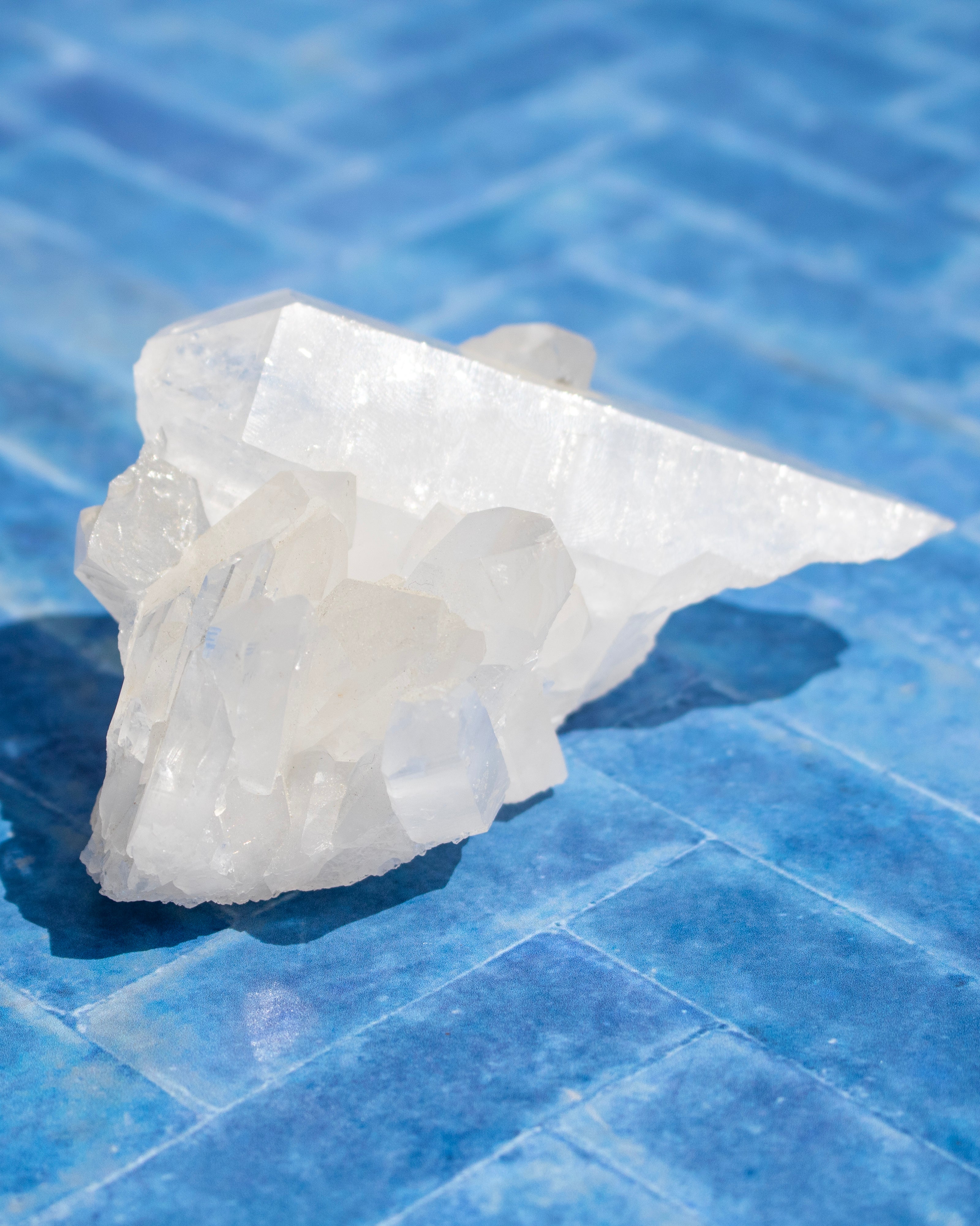 A raw, translucent white crystal cluster with sharp geometric facets, resting on a blue tiled surface.