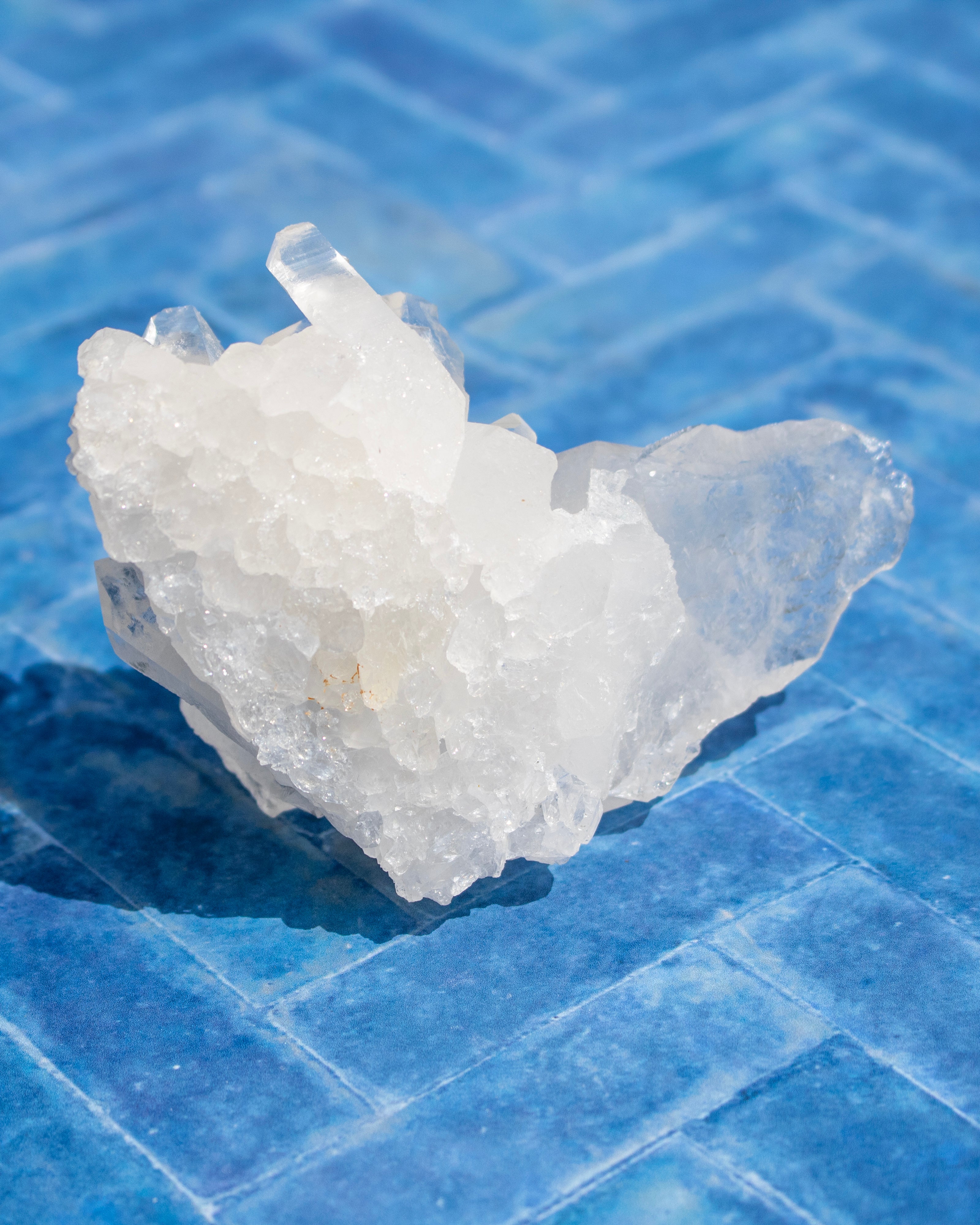 A raw white crystal cluster with rough, jagged edges and clear facets, resting on a blue tiled surface.