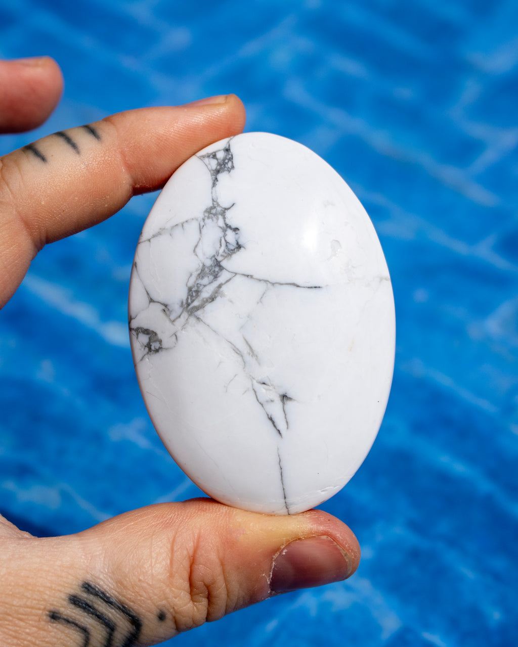 A hand holding a polished white oval stone with natural gray and black veining, set against a bright blue textured background.
