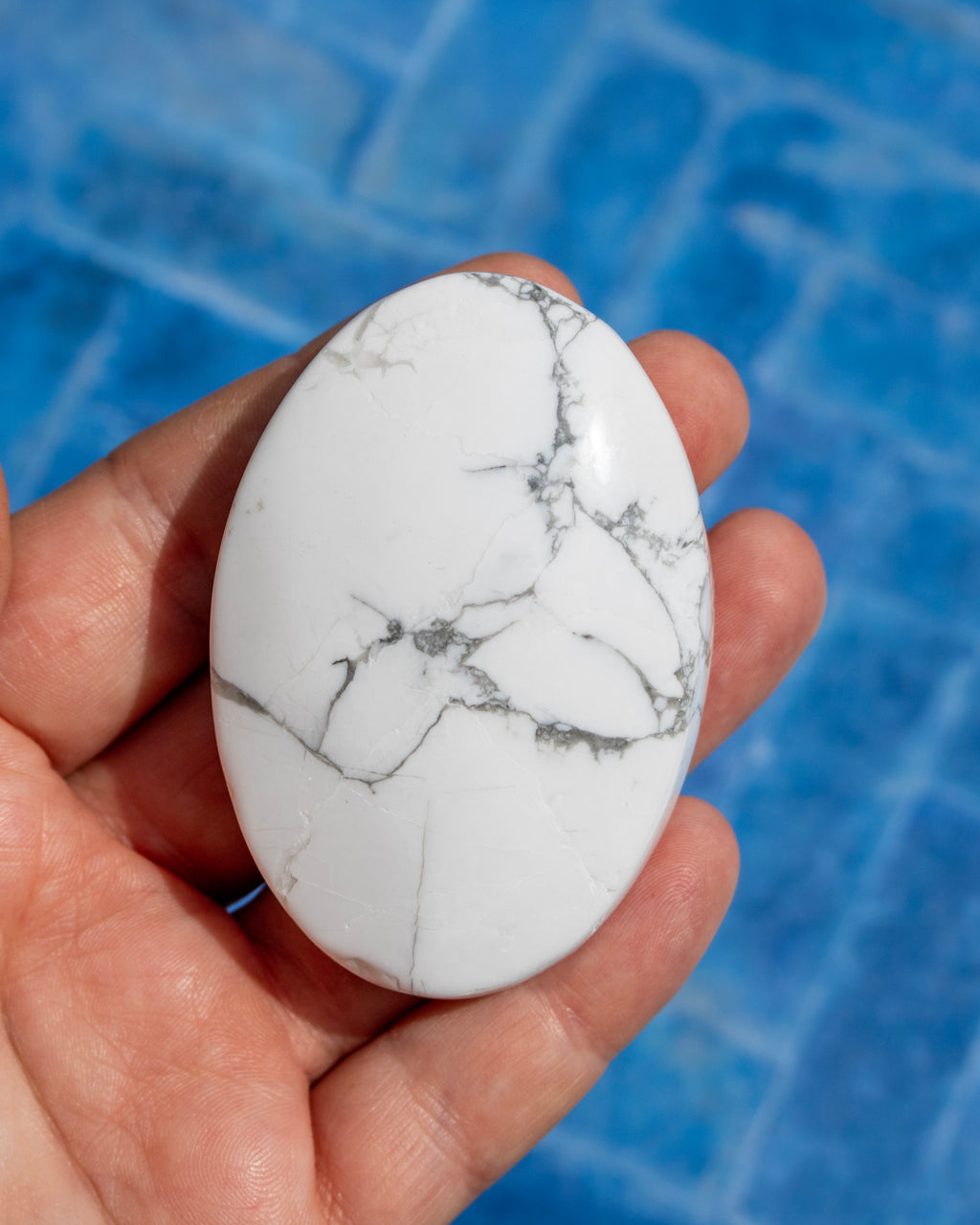 A hand holding a polished white oval stone with gray veining, set against a blue tiled background.