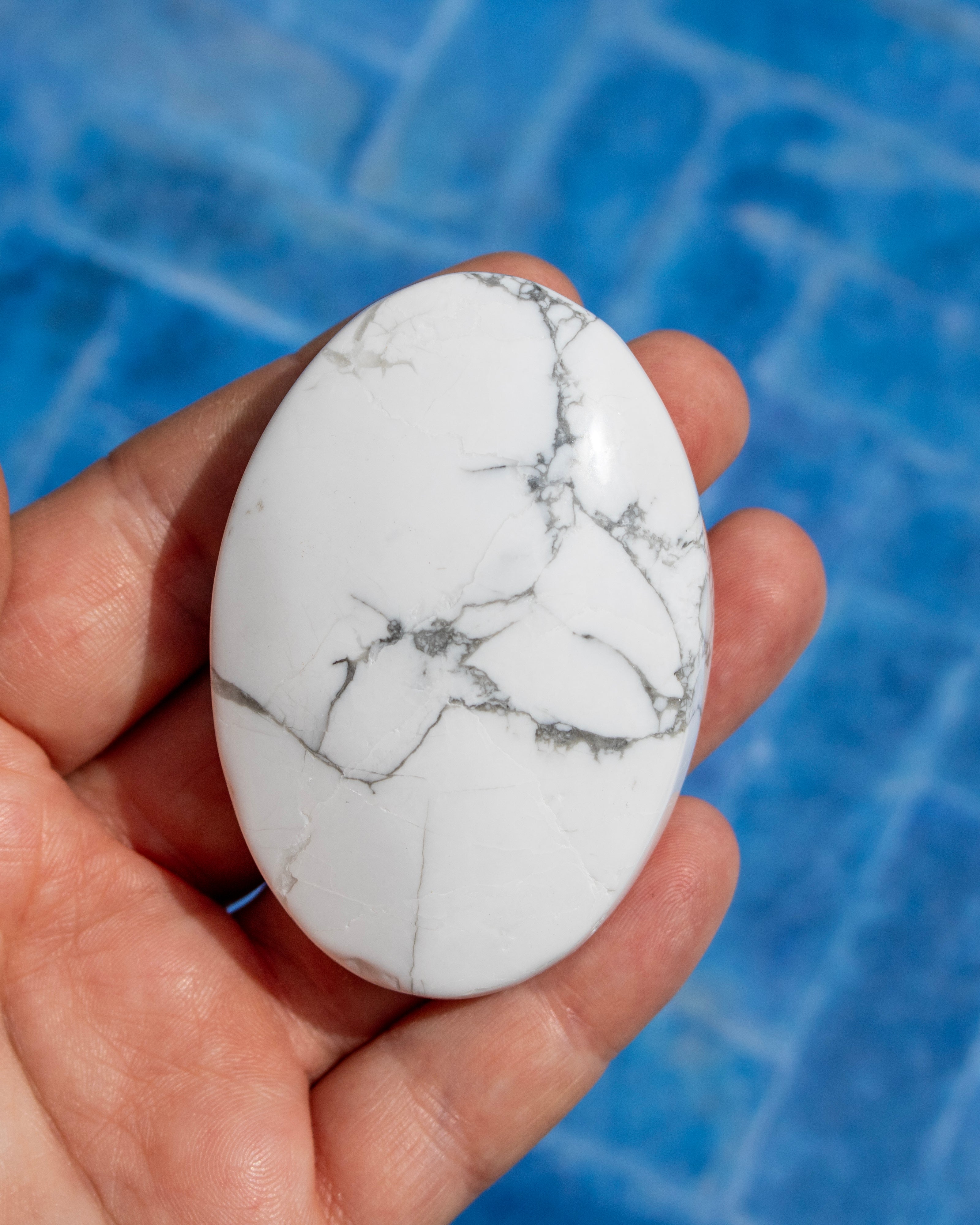 A hand holding a polished white oval stone with gray veining, set against a blue tiled background.